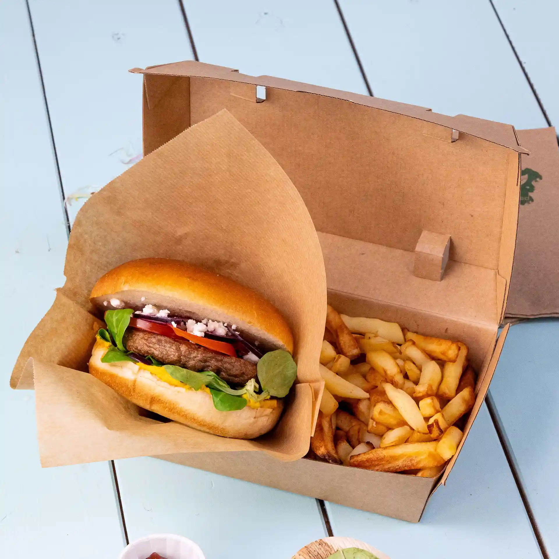 Brown cardboard food box containing a hamburger with toppings and a side of fries, placed on a light blue surface.