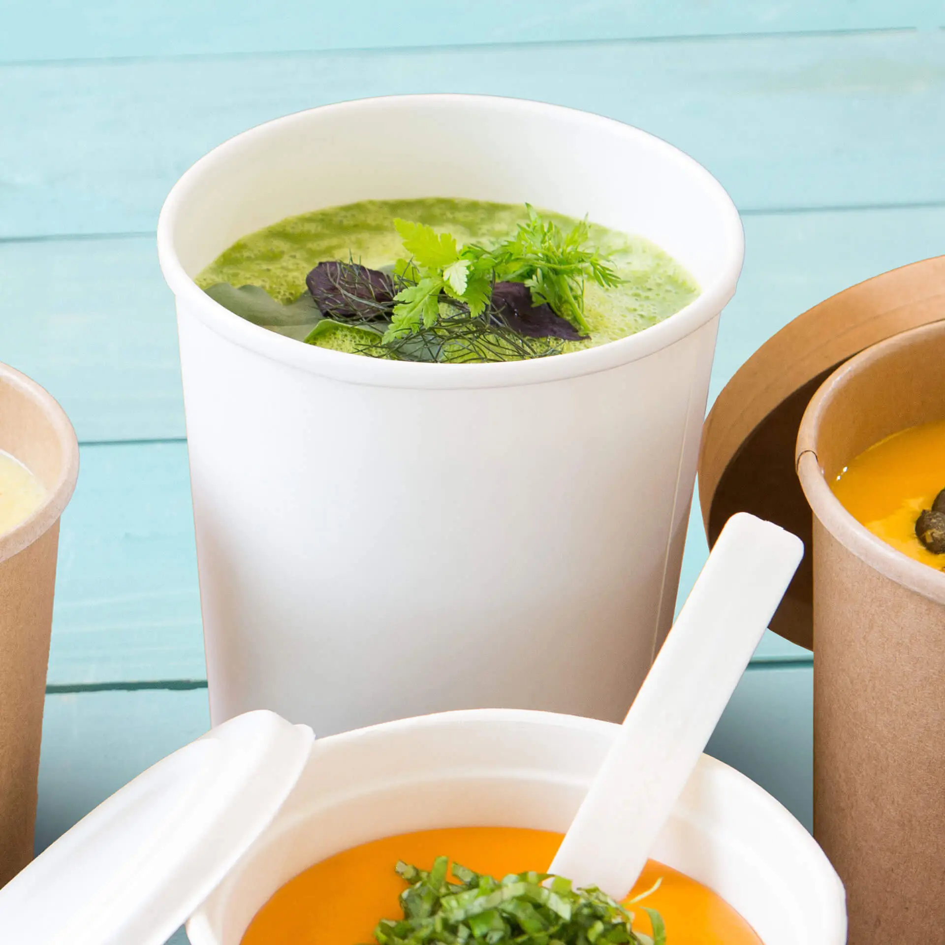 White biodegradable soup cup with green soup and herbs, positioned with other soup containers on a teal background.