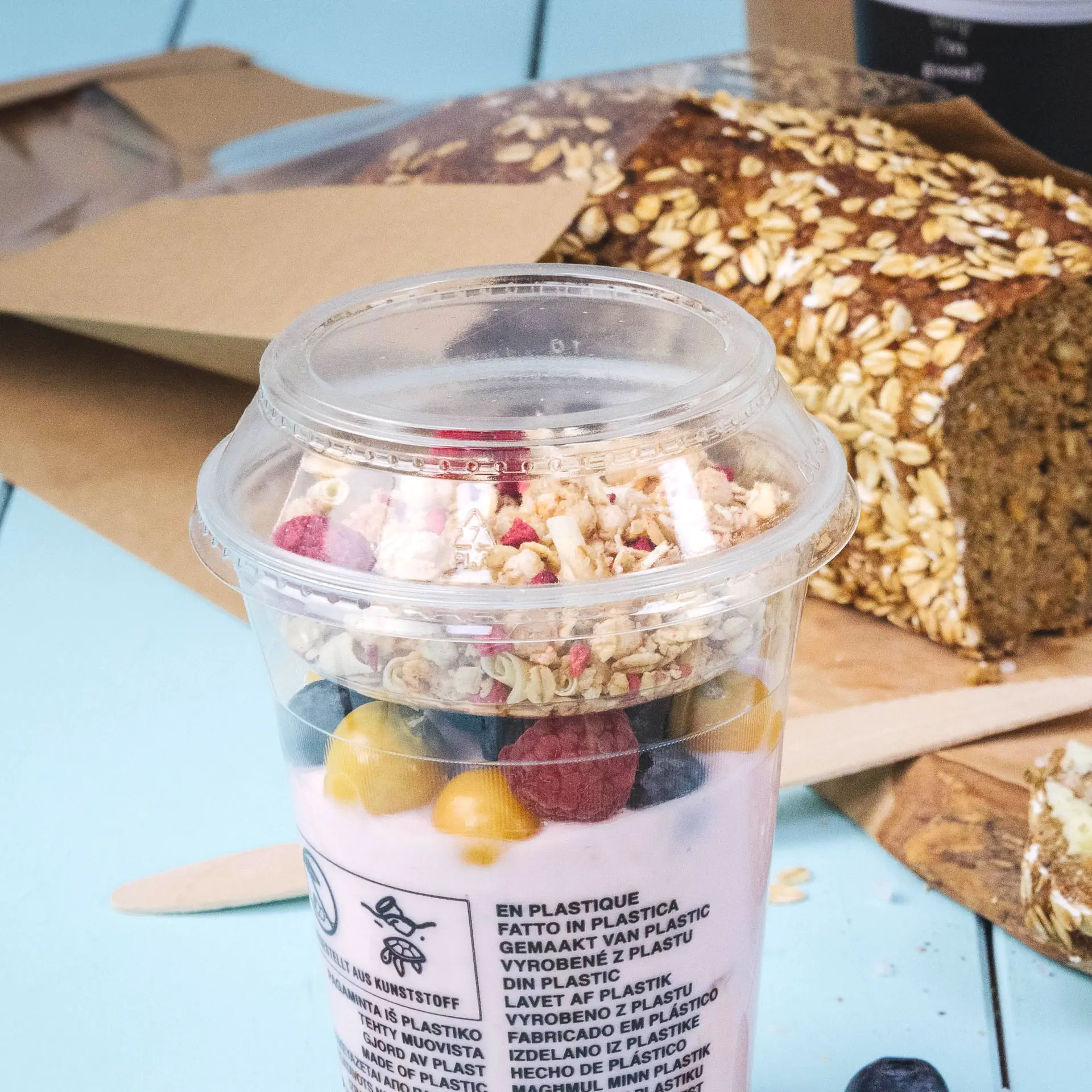 Clear PLA combo lid with granola, berries, and yogurt inside; oat bread in the background; eco-friendly design noted.