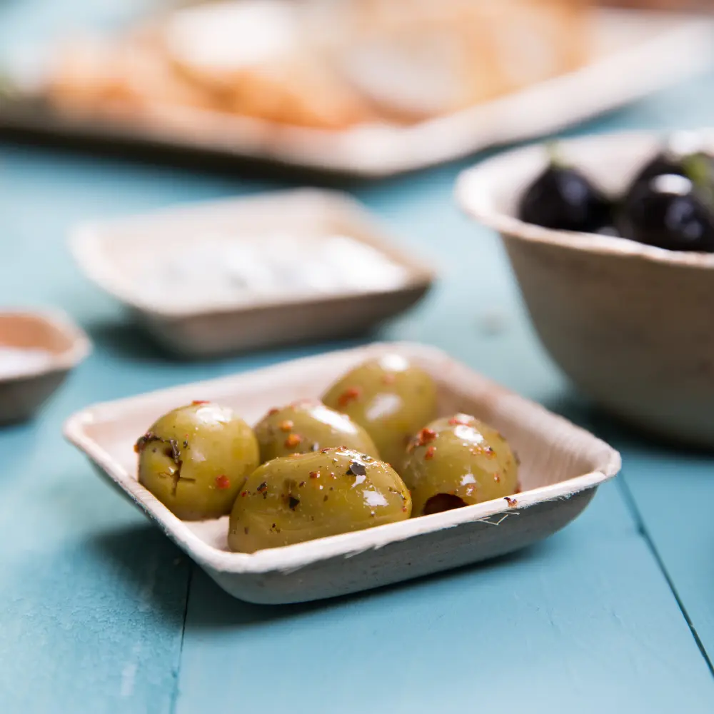 A square disposable bowl containing several green olives with red seasoning, placed on a blue wooden surface.