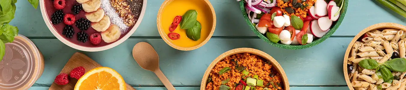 Colorful bowls filled with various foods, including fruit, pasta, salad, and grains, arranged on a wooden table.