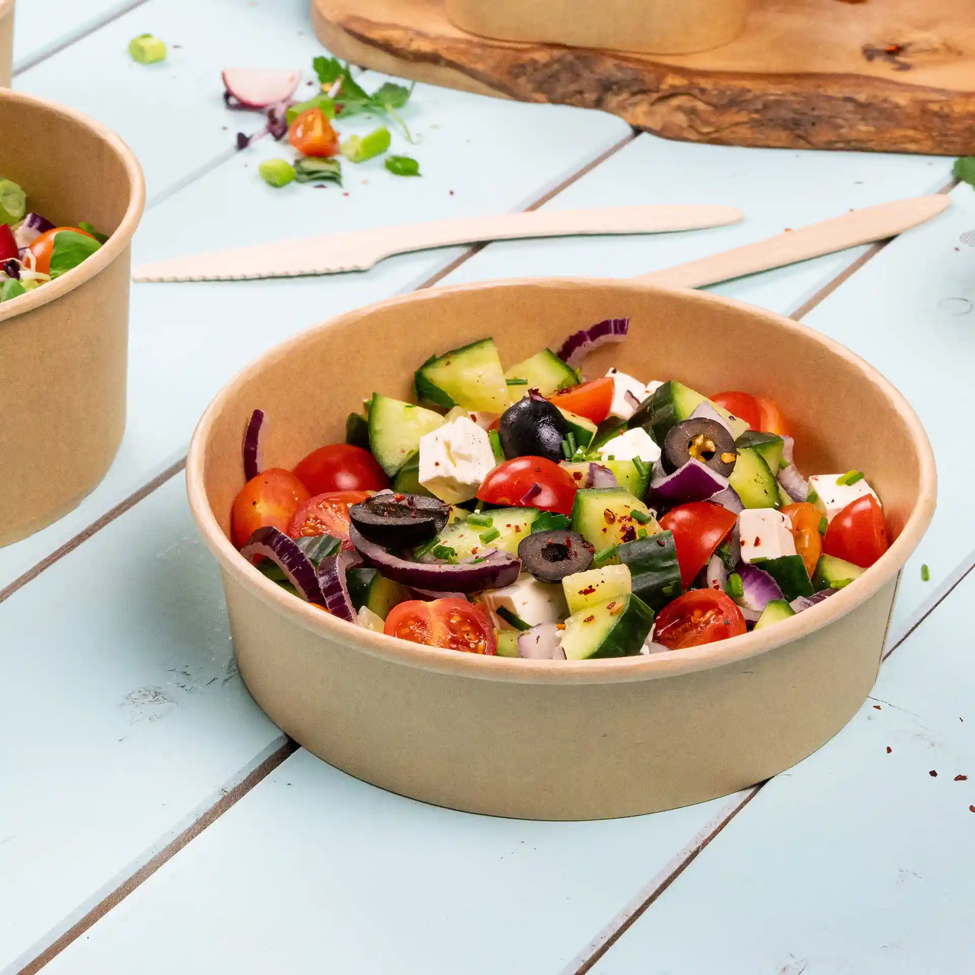 Round brown cardboard bowl filled with salad, including tomatoes, cucumbers, olives, and feta cheese, on a blue surface.