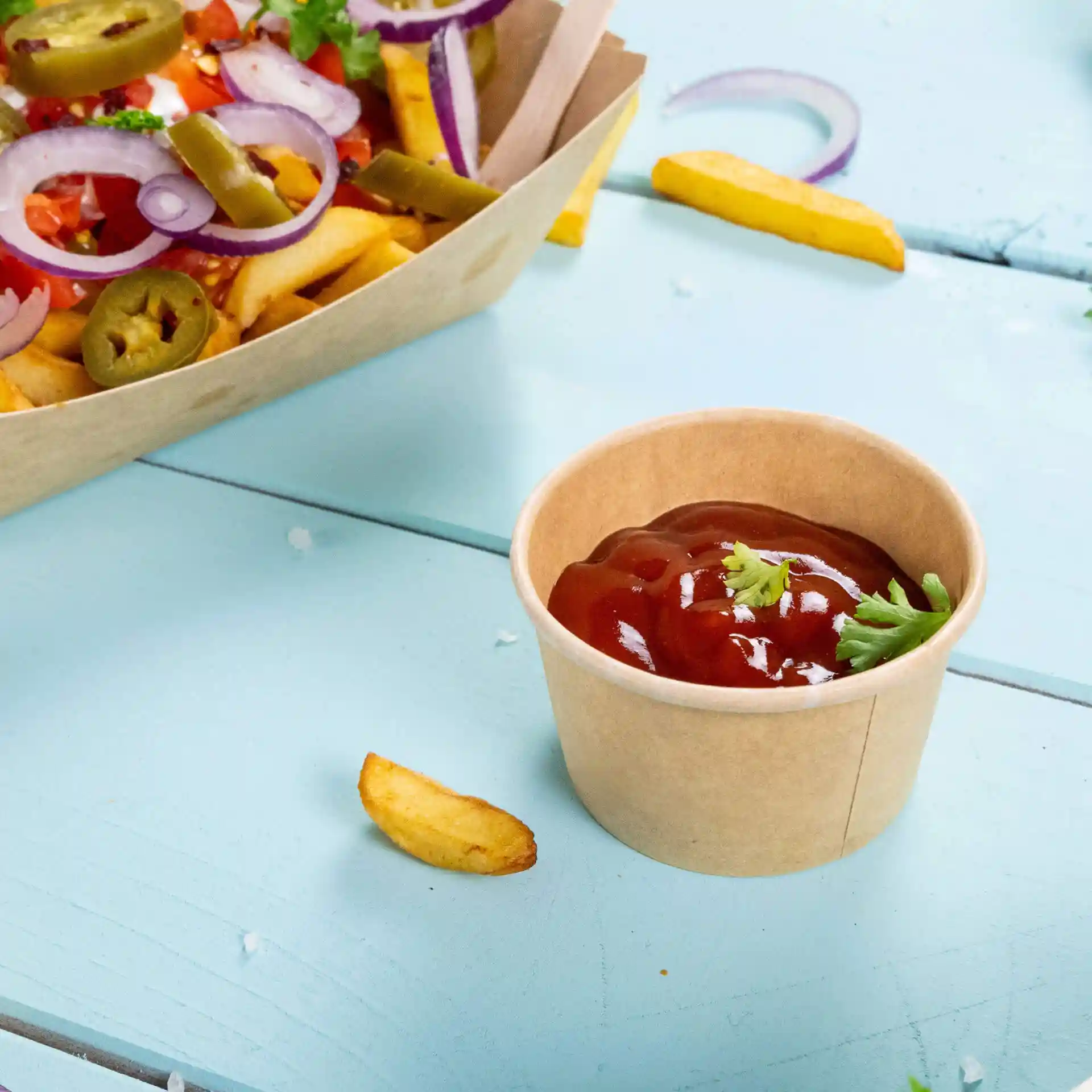 Brown paper portion cup with ketchup and a garnish of cilantro, placed on a light blue surface with fries nearby.