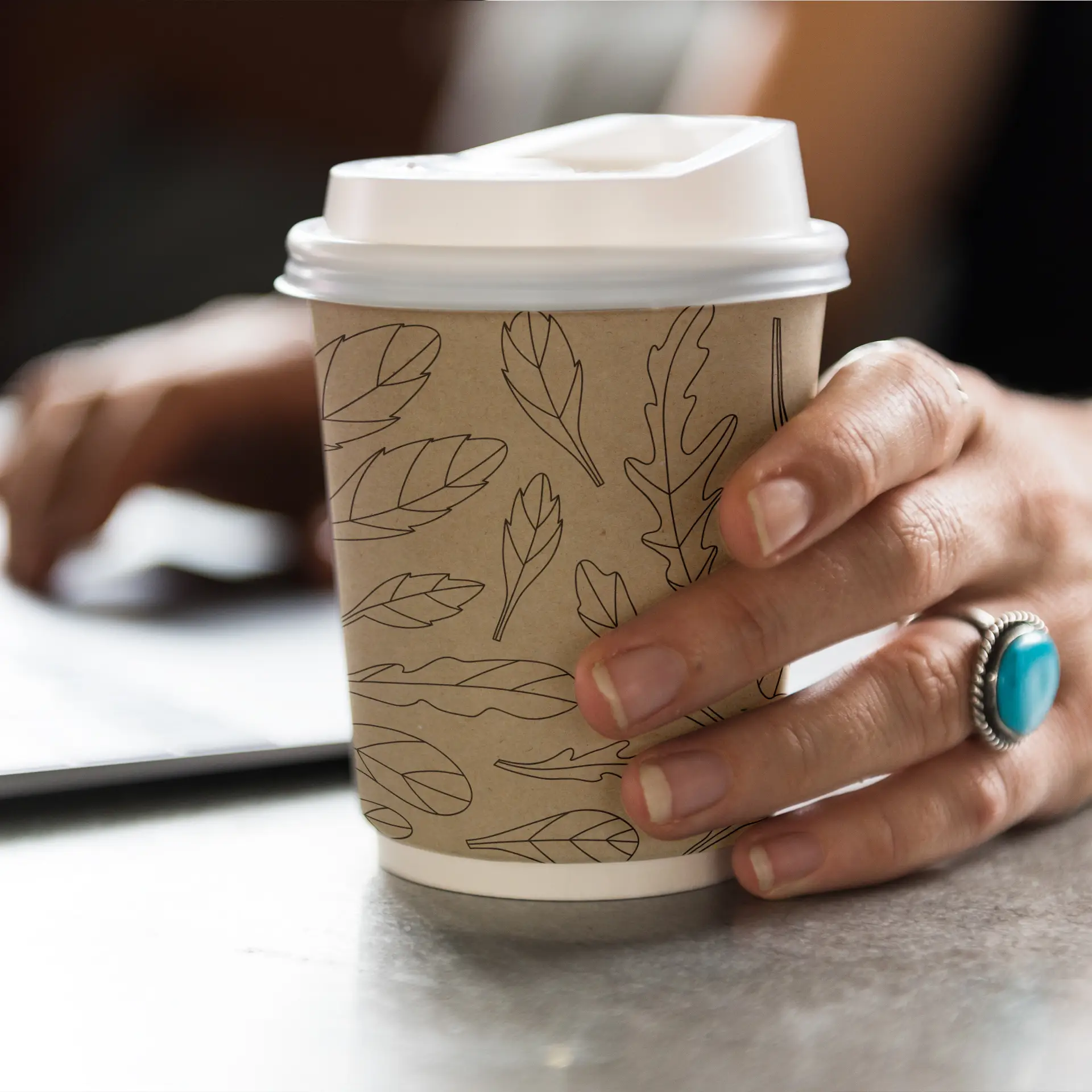 A hand holding a double-wall paper cup featuring a leaf design; a blurred hand is reaching for a laptop in the background.