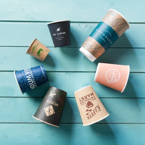 Various disposable coffee cups with unique designs scattered on a turquoise wooden surface.