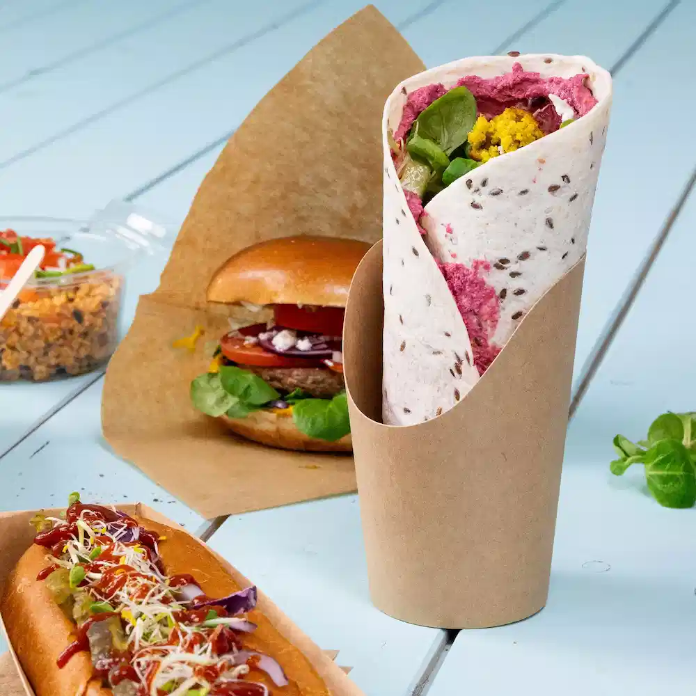 Wrap filled with greens and spread, served in a brown paper holder, with a burger and hot dog in the background.