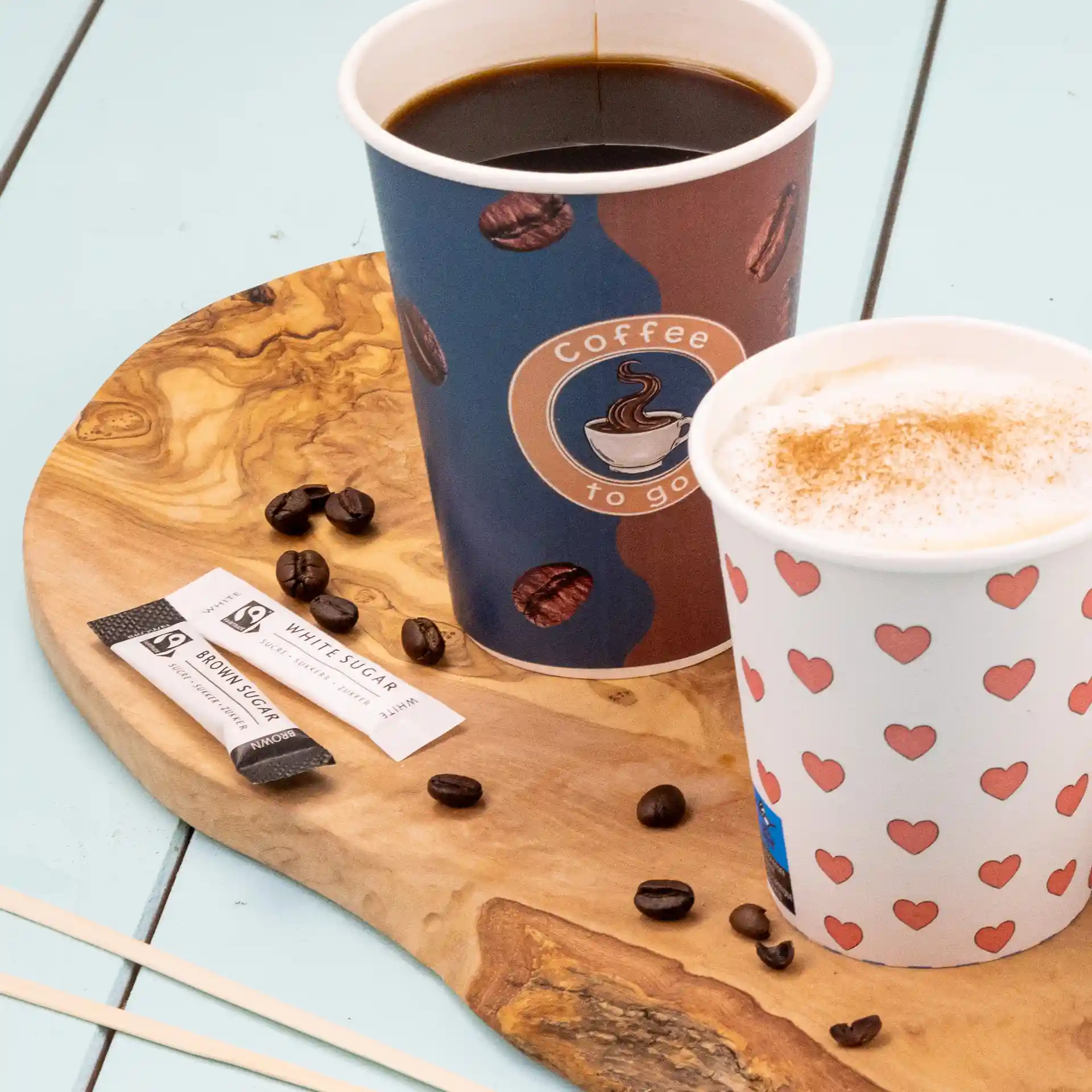 Two coffee cups on a wooden serving board with sugar sticks and coffee beans scattered around.