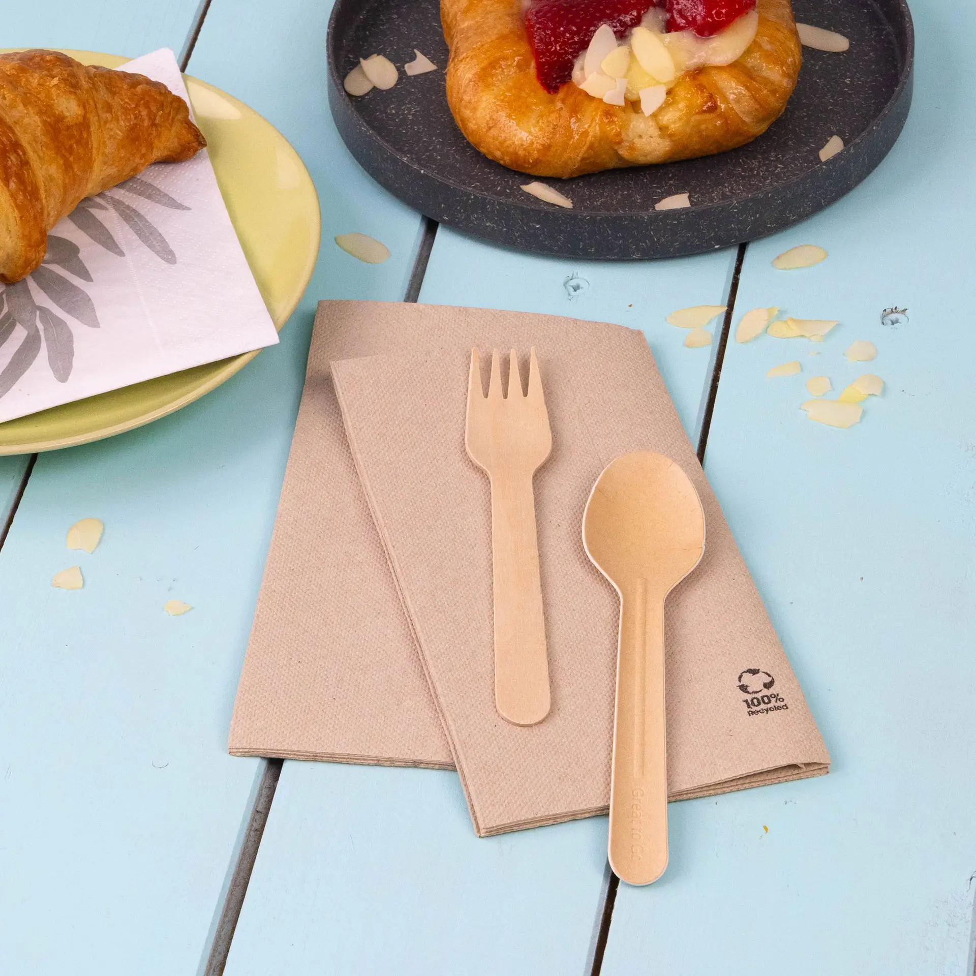 Brown recycled paper napkins folded with a biodegradable spoon and fork on a blue surface near a croissant and a pastry.