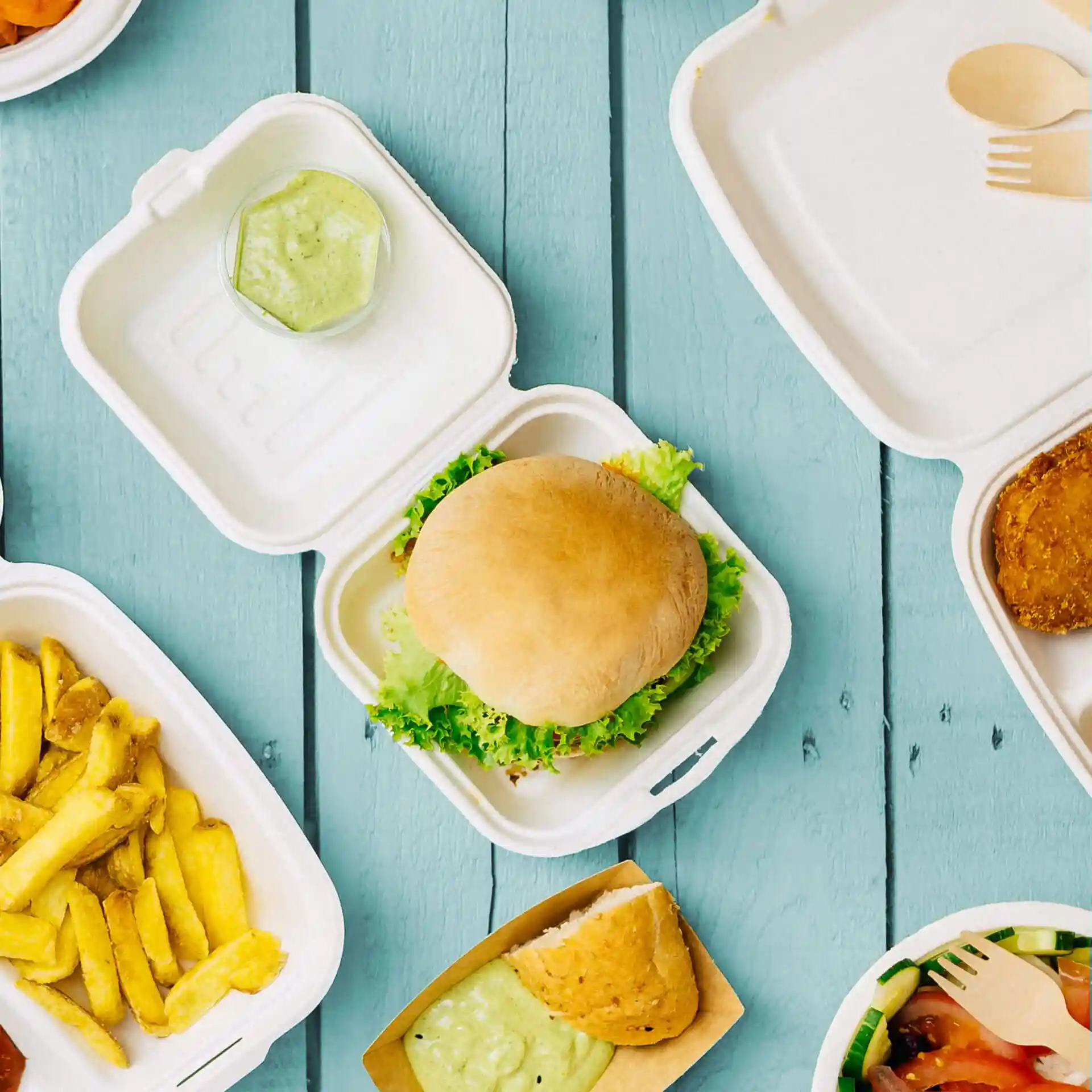 Image of a sugarcane burger box containing a burger with lettuce, a small container of sauce, and various food items nearby.