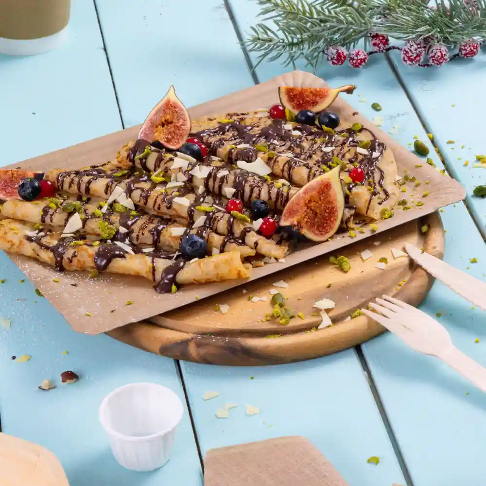 A wooden platter with rolled crepes drizzled with chocolate, garnished with figs, berries, and pistachios. Fork and knife beside.