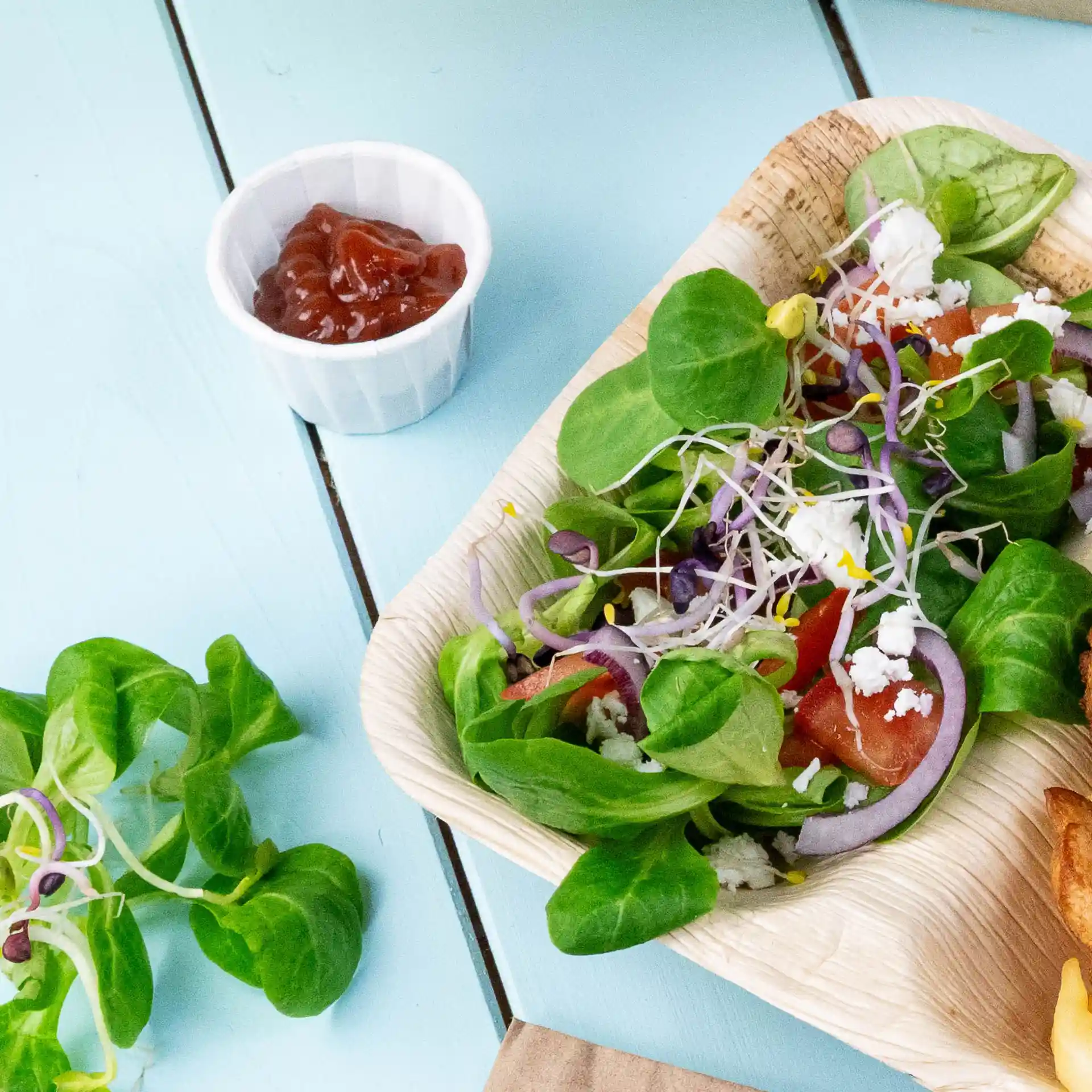 Salad with mixed greens, tomatoes, onions, and sprouts in a palm leaf bowl, accompanied by a small white cup of ketchup.