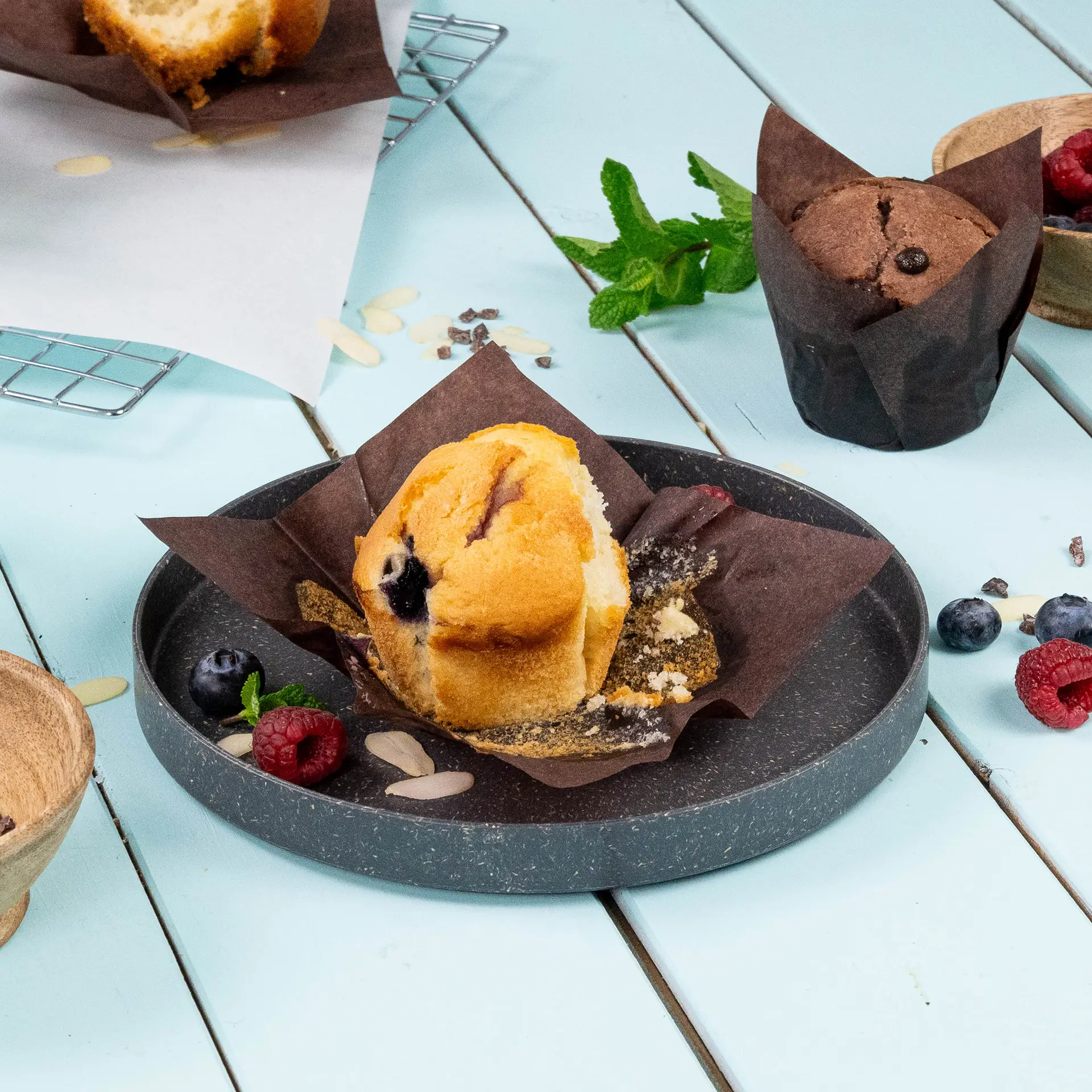 A brown paper muffin cup with a blueberry muffin sitting on a dark plate, surrounded by raspberries, blueberries, and a mint leaf.