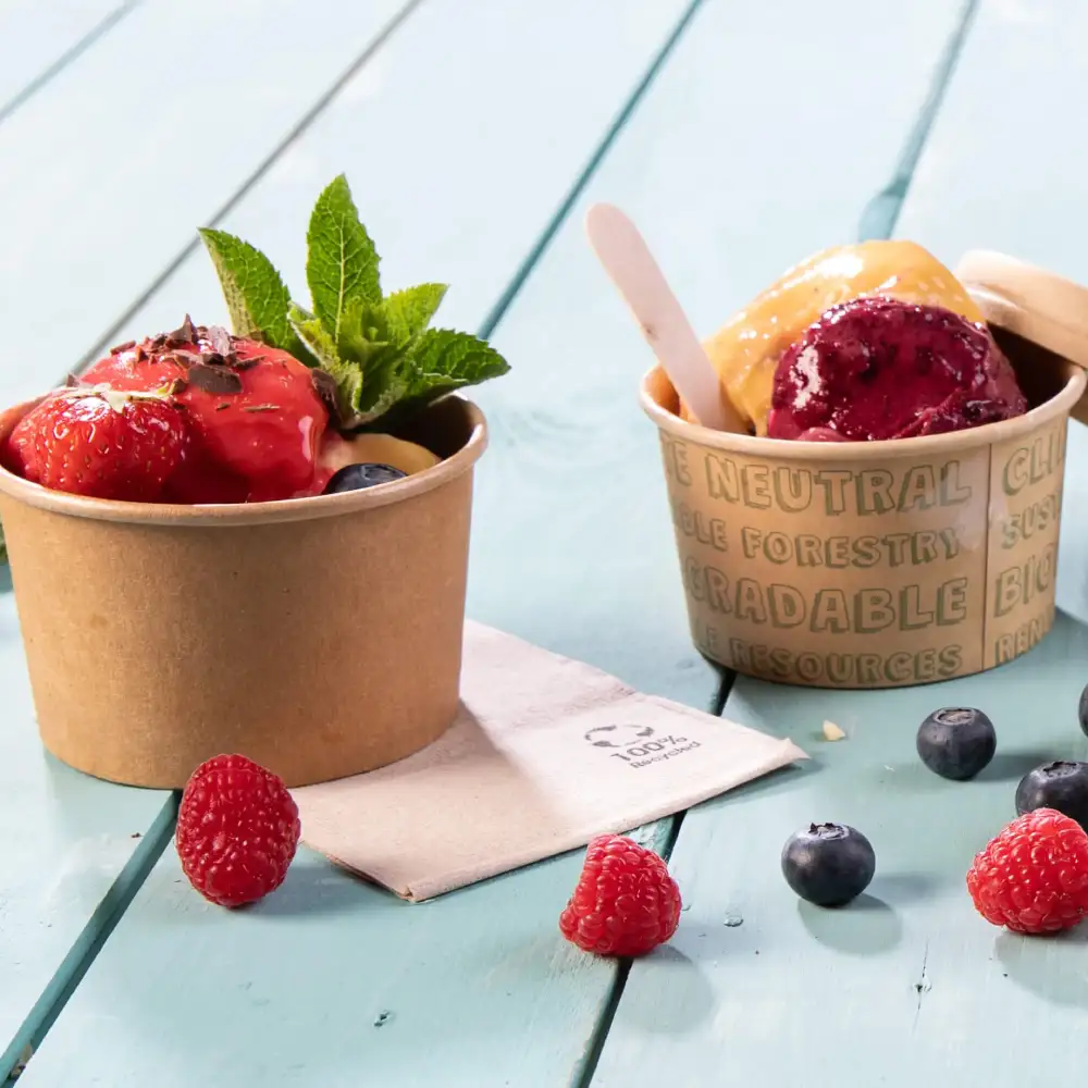 Two ice cream cups with berries on a blue wooden surface, one topped with strawberries and mint, the other with purple and yellow ice cream.