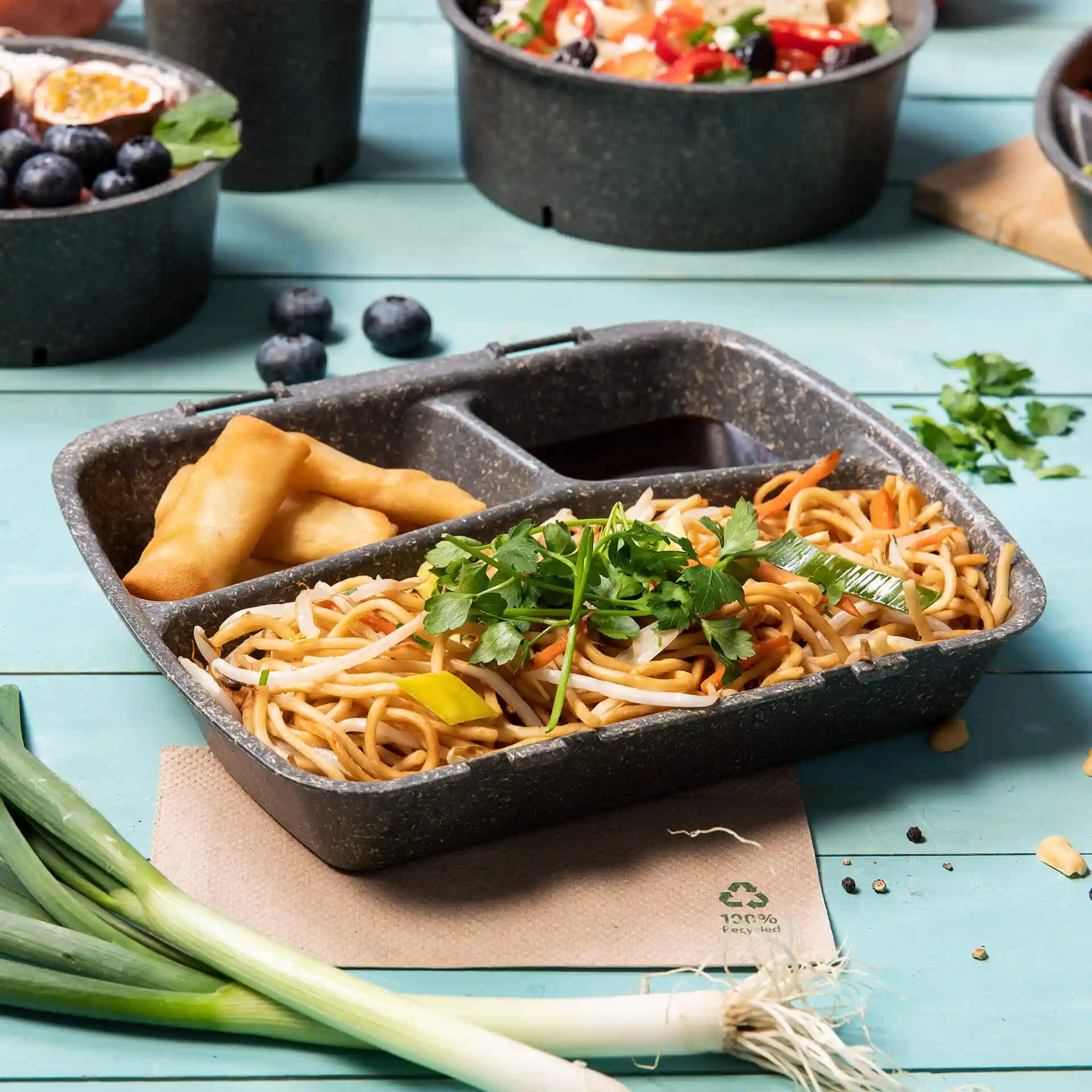 Reusable grey meal container with three compartments, filled with noodles, sauce, and spring rolls, on a blue surface.
