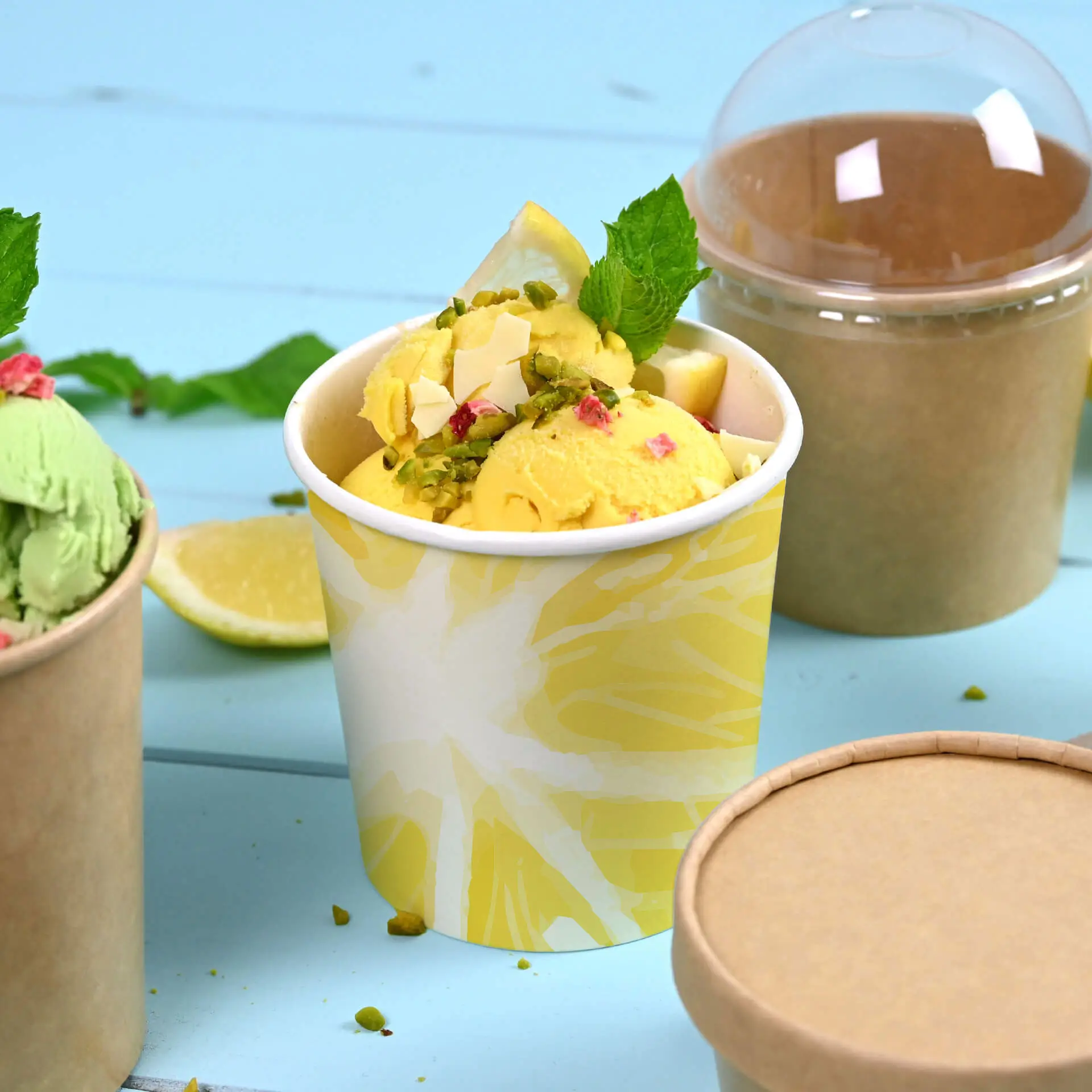 Printed ice cream cup with yellow design, filled with ice cream, mint leaf, and toppings, next to other cups on a blue background.