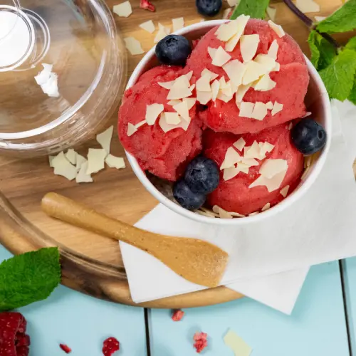 Bowl of raspberry sorbet topped with blueberries and white chocolate shavings, wooden spoon, and mint leaves on a wooden surface.