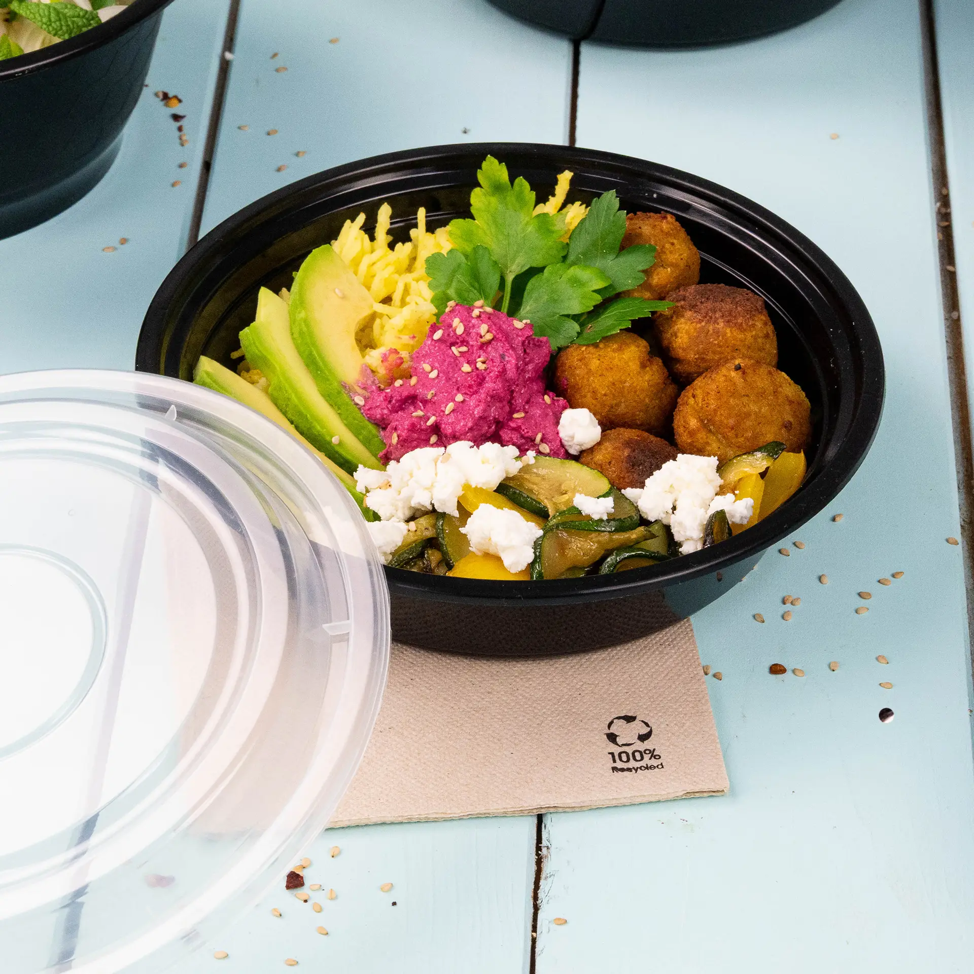 A black reusable plate filled with colorful ingredients including avocado, hummus, and falafel balls, with a clear lid partially removed.