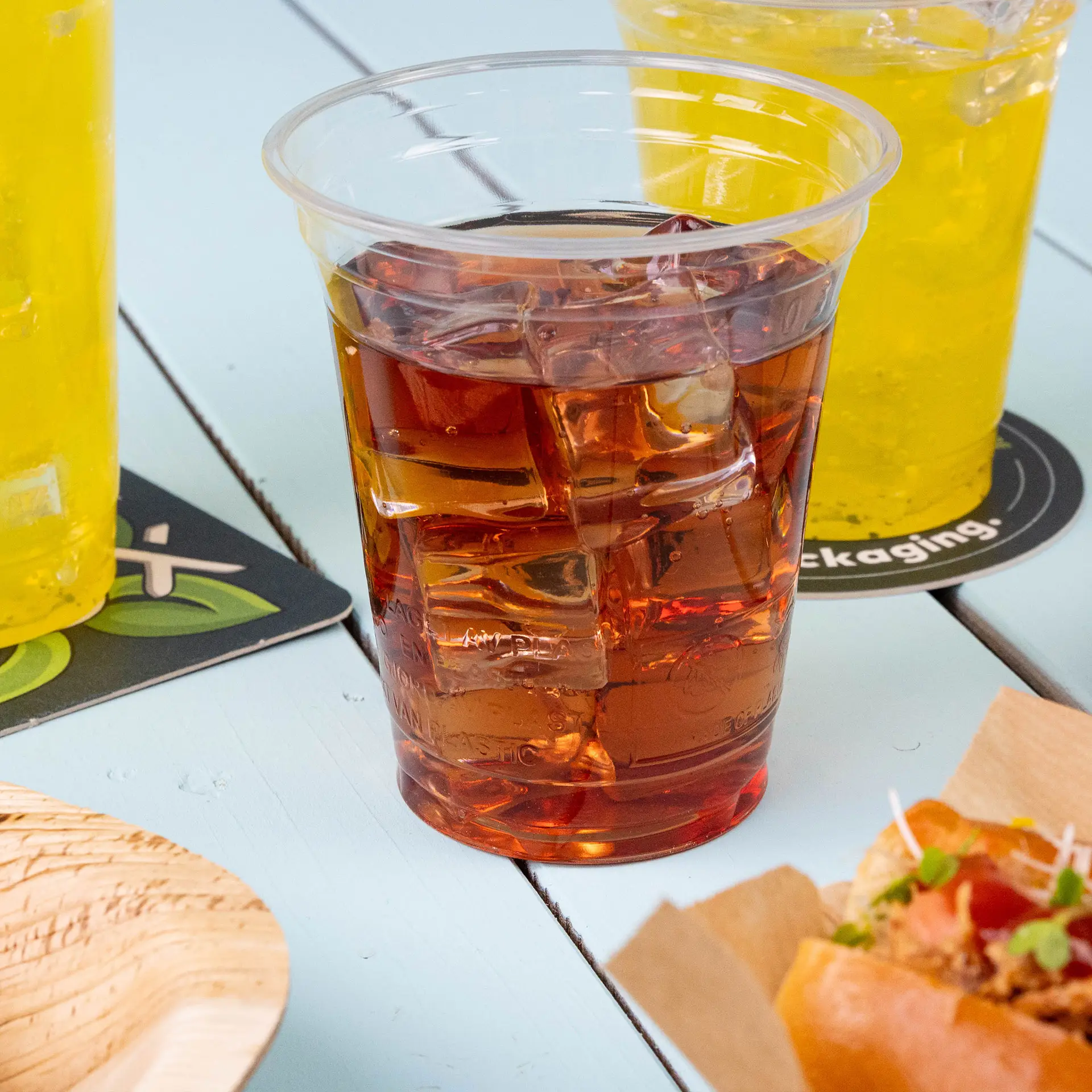 Clear plastic cup filled with dark liquid and ice, surrounded by two yellow drinks and a wooden plate on a light blue table.