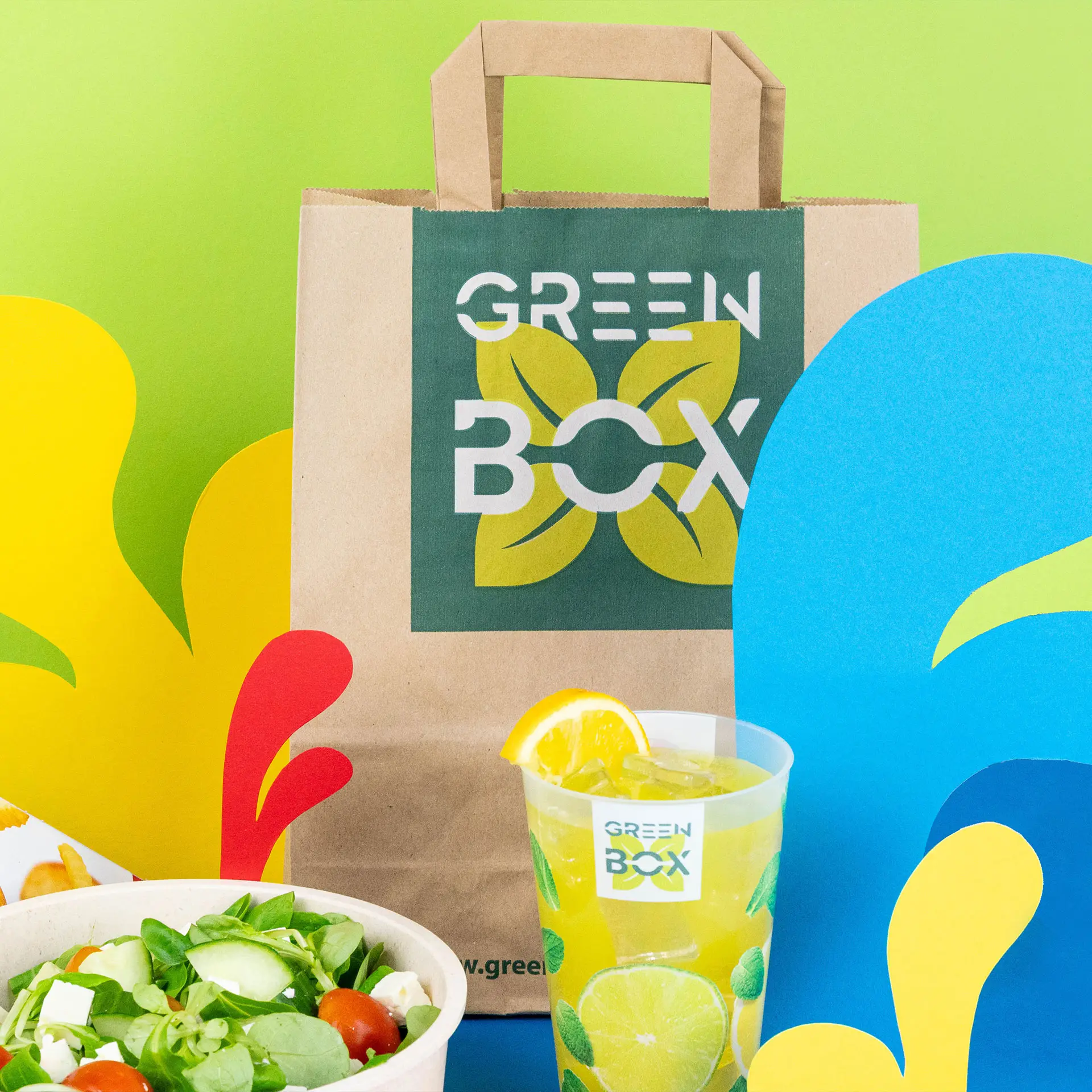 Brown paper carrier bag with a green logo, a salad bowl, and a drink with a lemon slice on a colorful background.