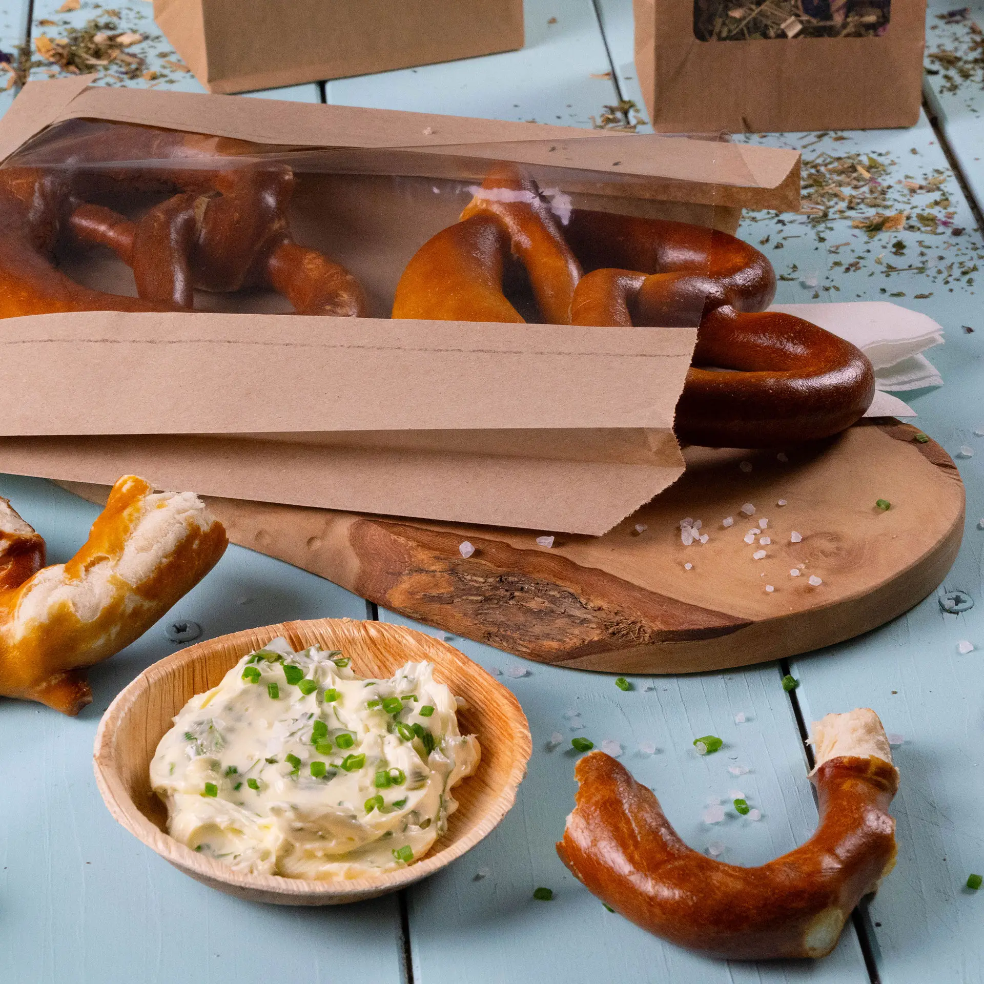 Brown paper bread bag with window, containing soft pretzels and a bowl of creamy dip topped with chives on a wooden board.