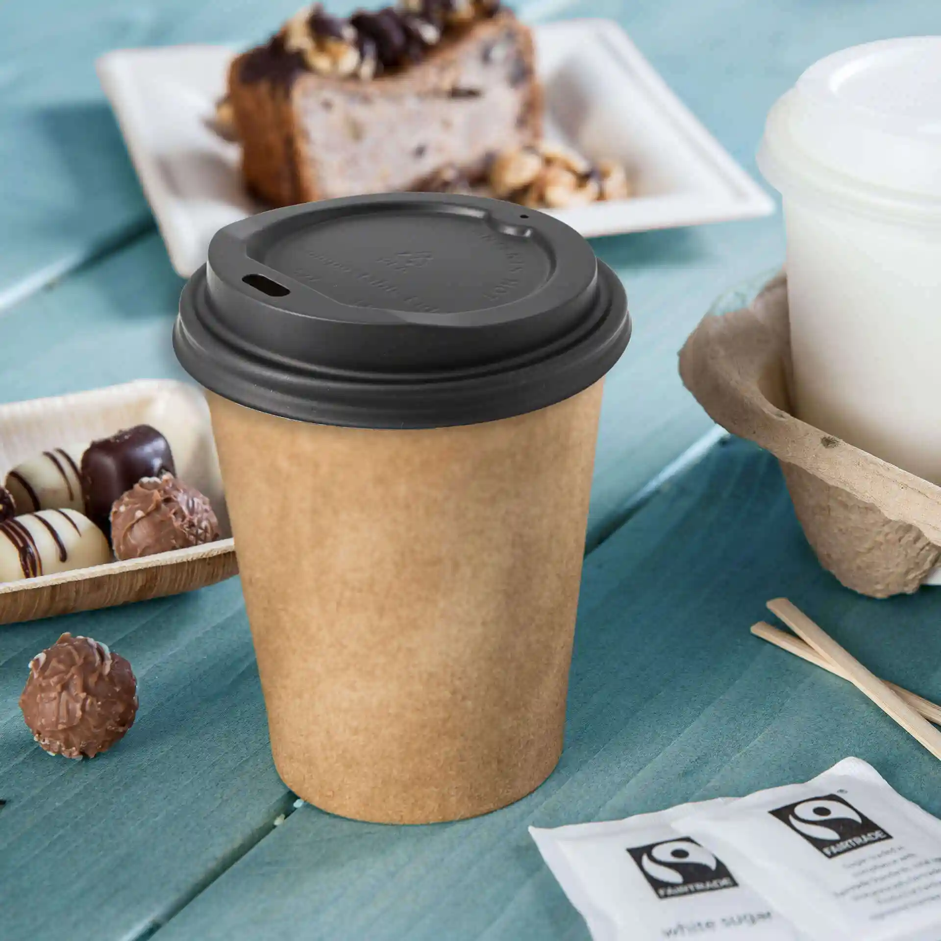 Brown paper cup with a black lid, surrounded by chocolates, sugar packets, and a dessert on a blue wooden surface.