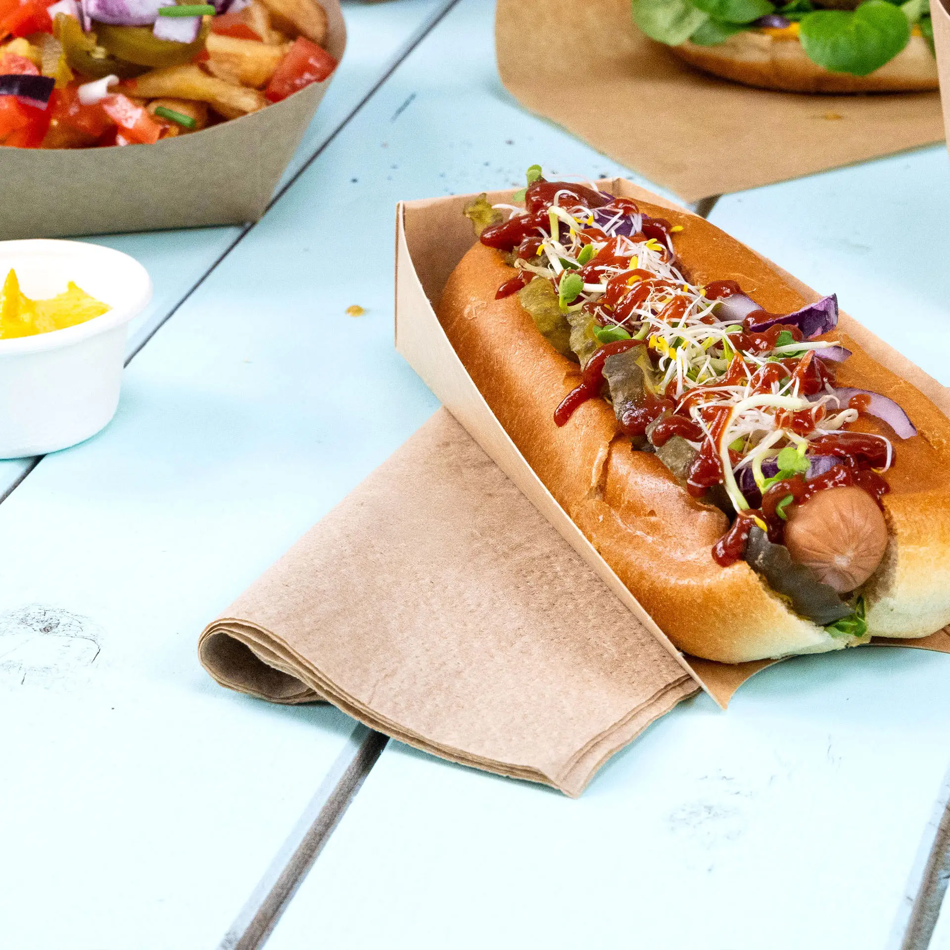 Hot dog in a paper holder topped with ketchup, mustard, pickles, and sprouts, with a square unbleached napkin beside it.