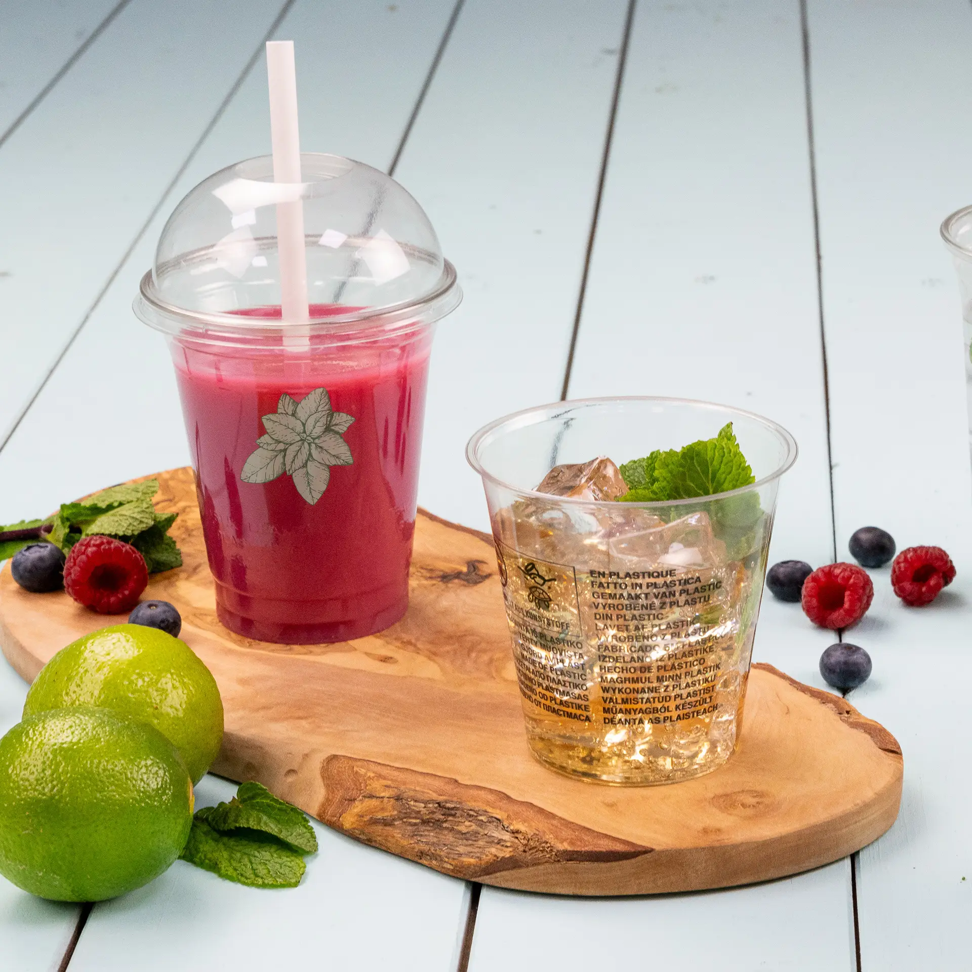 Clear rPET cup for cold drinks, featuring a leafy design, with a second cup beside it on a wooden board; garnished with mint and lime.