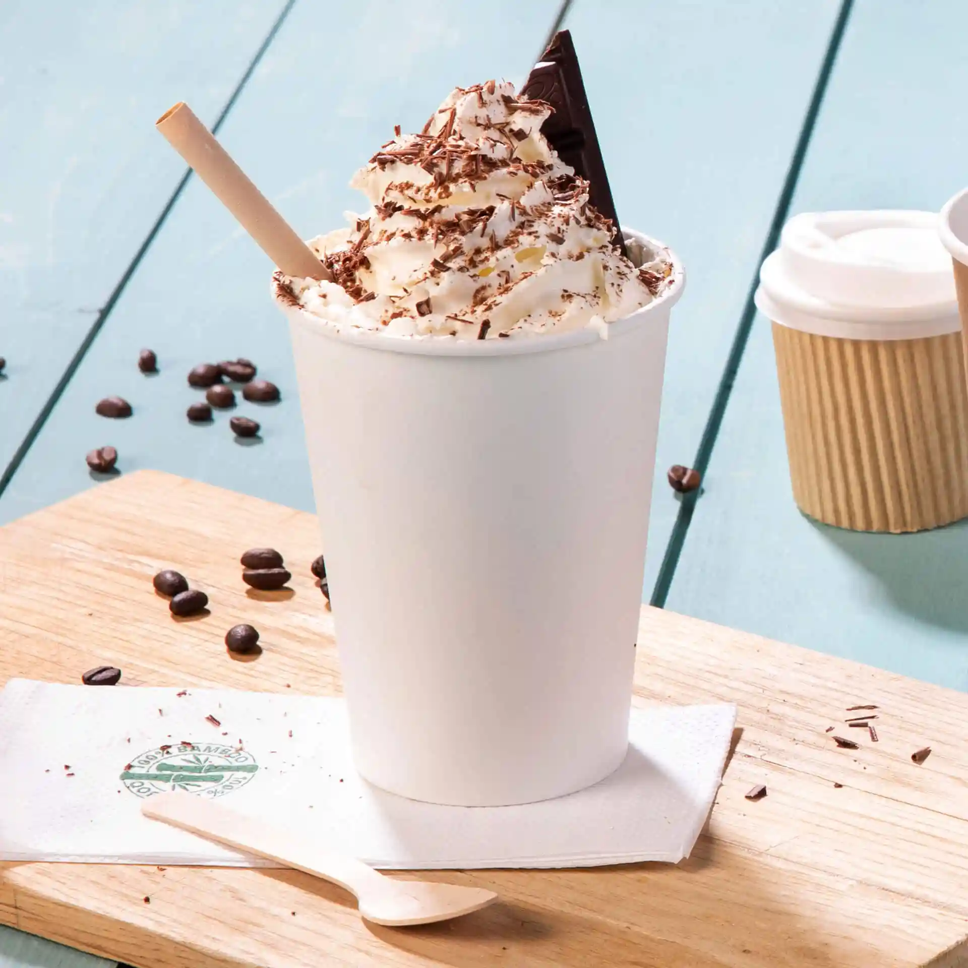 White paper cup filled with whipped cream and chocolate shavings, with a wooden spoon beside it on a wooden surface.