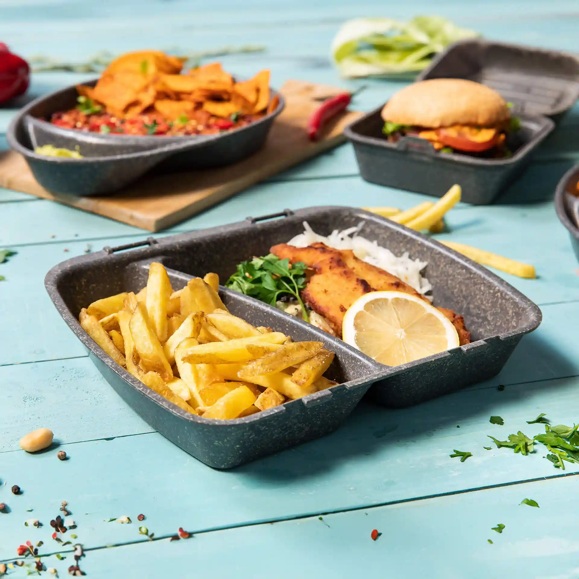 Reusable grey meal container with two compartments, filled with fries and fish, lemon slice, and garnished with herbs.