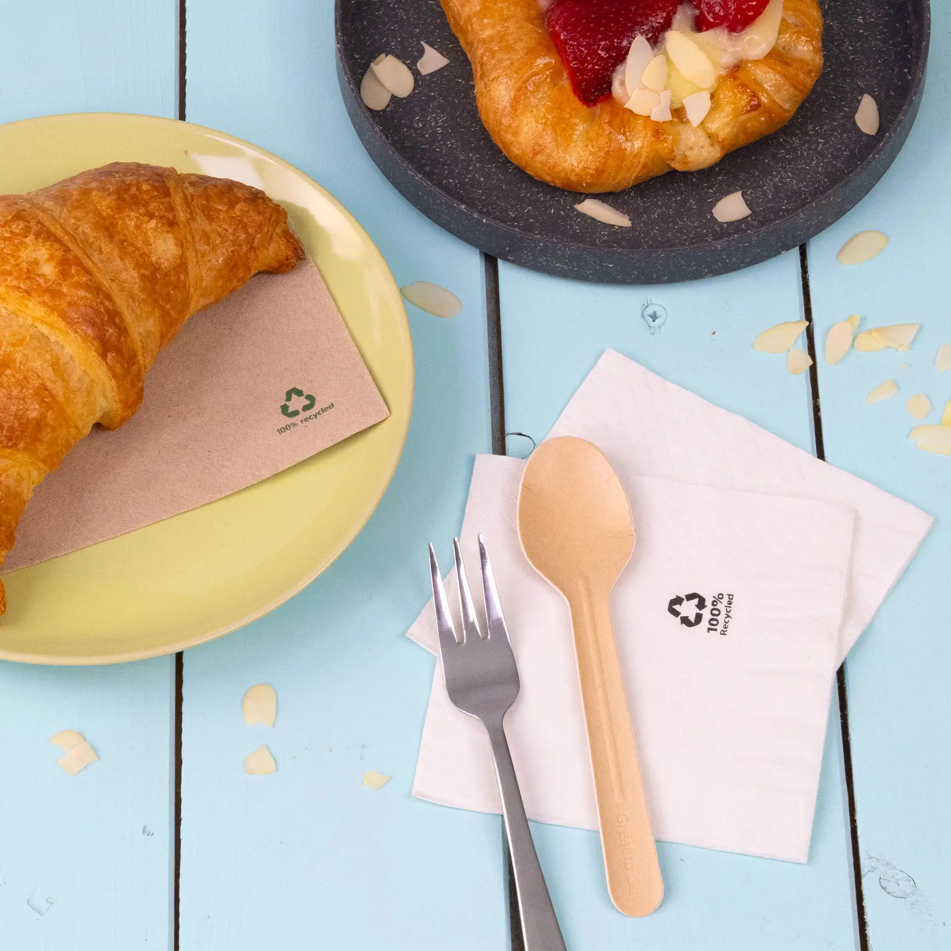 A croissant on a yellow plate beside a white 2-ply recycled paper napkin and an eco-friendly wooden spoon and fork.