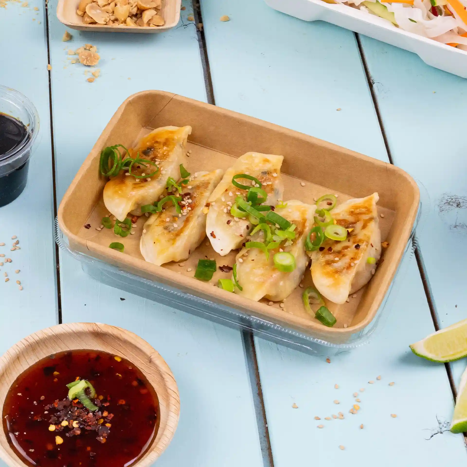 Container with five dumplings topped with green onions and sesame seeds, served with a dipping sauce in a wooden bowl.