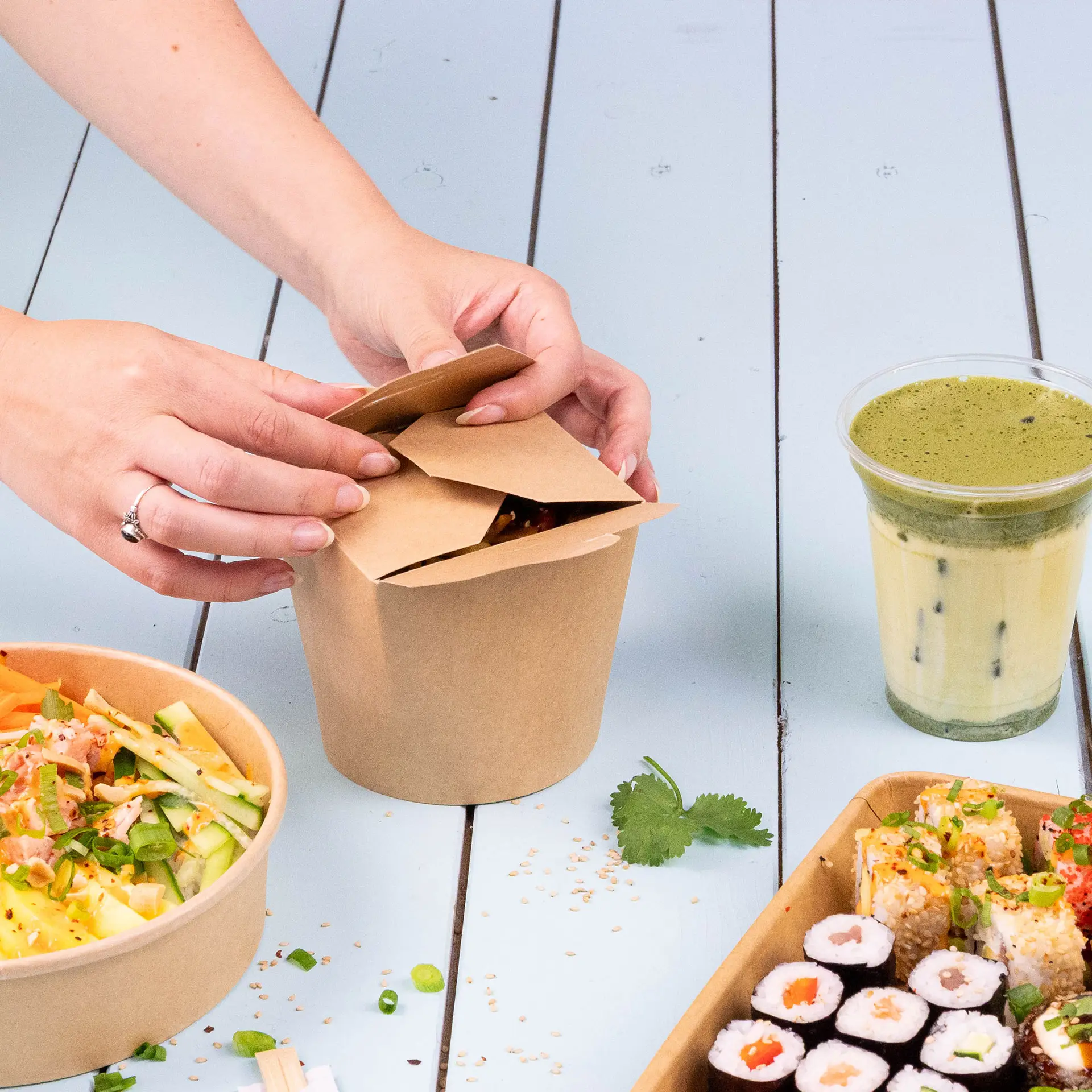 Noodle box made of kraft paperboard, filled with fried noodles, topped with tomato slices and cilantro, on a bamboo mat.