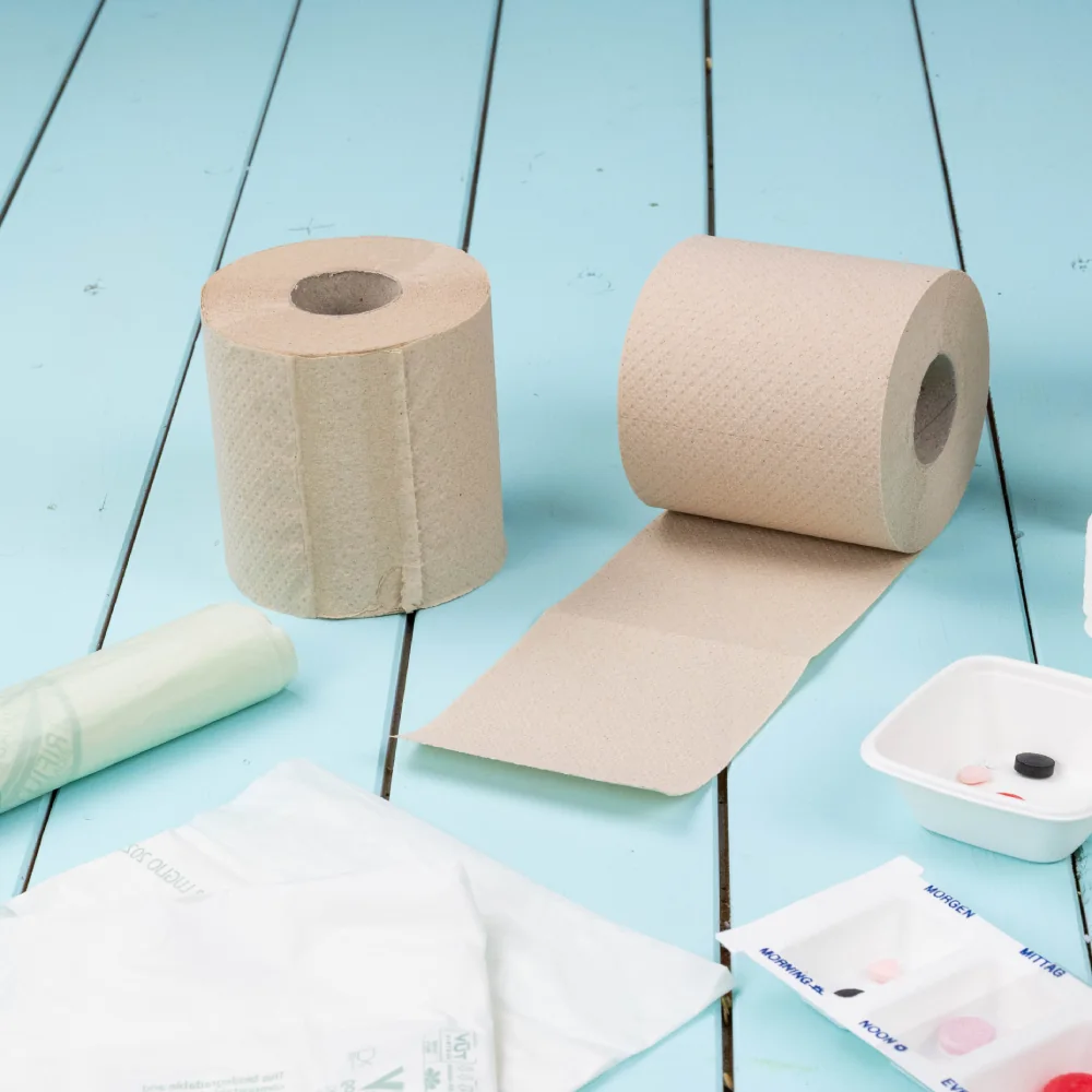 Roll of brown paper towels next to another roll with paper extending; additional items include a green plastic wrap, white bags, and a small dish with pills.