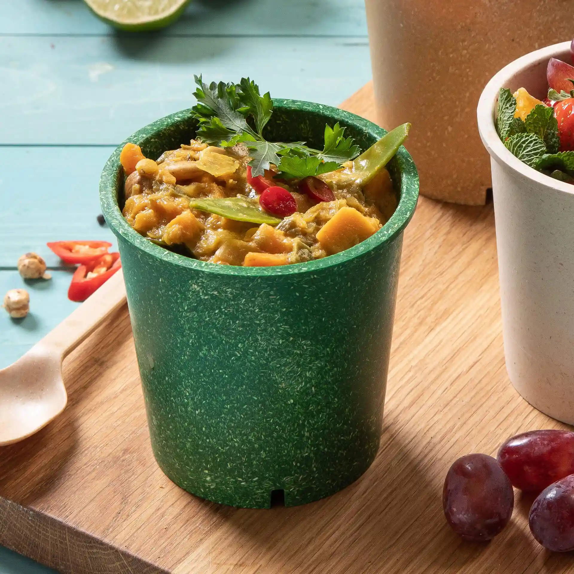 Green reusable cup filled with food, topped with cilantro and garnished with red chili and green beans.