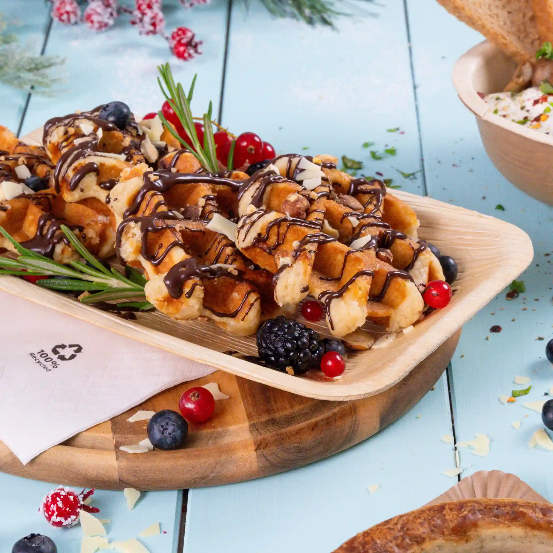 Waffles topped with chocolate drizzle, white chocolate shavings, and garnished with berries on a palm leaf plate.
