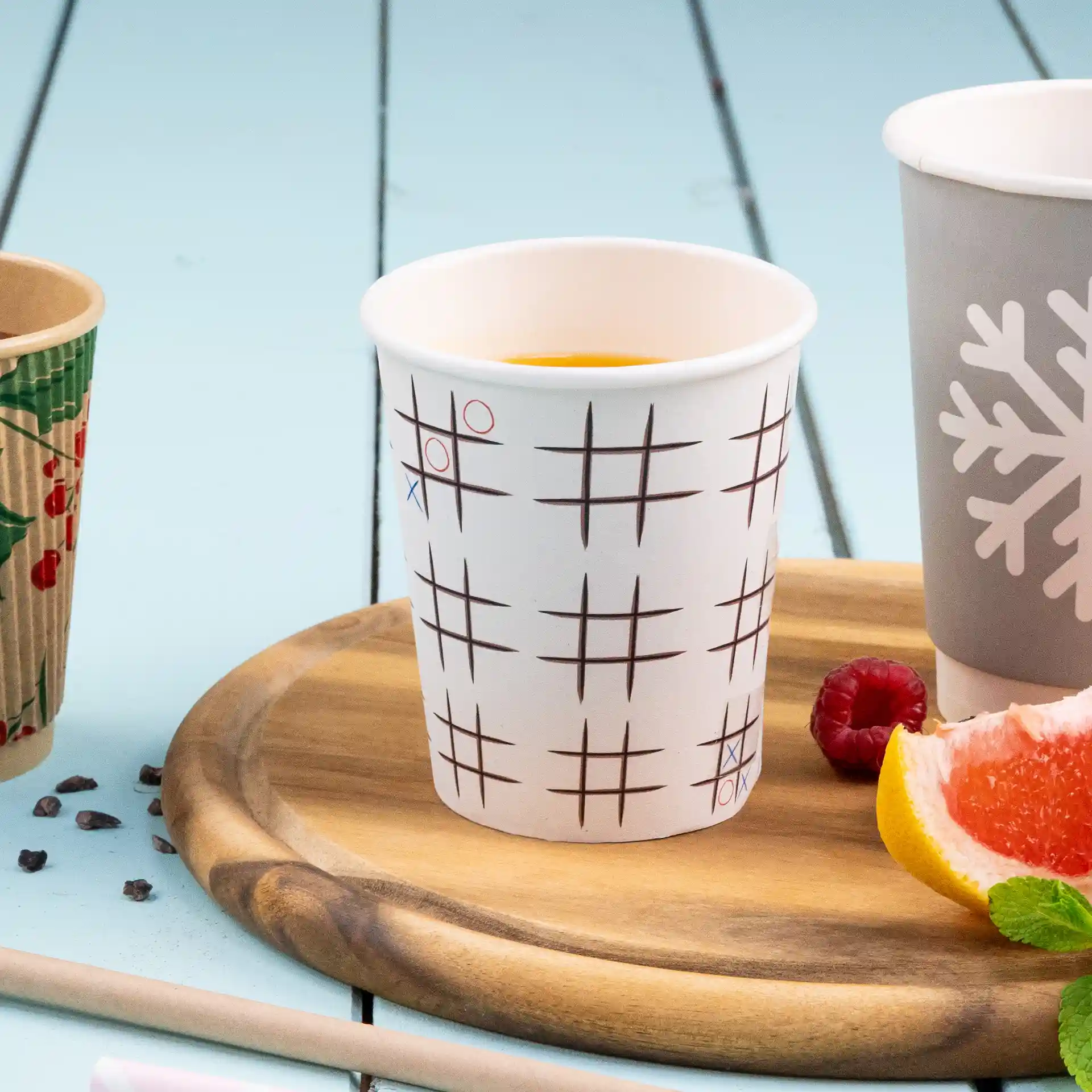 White paper cup with a tic-tac-toe pattern, placed on a wooden tray with fruit and another cup nearby.