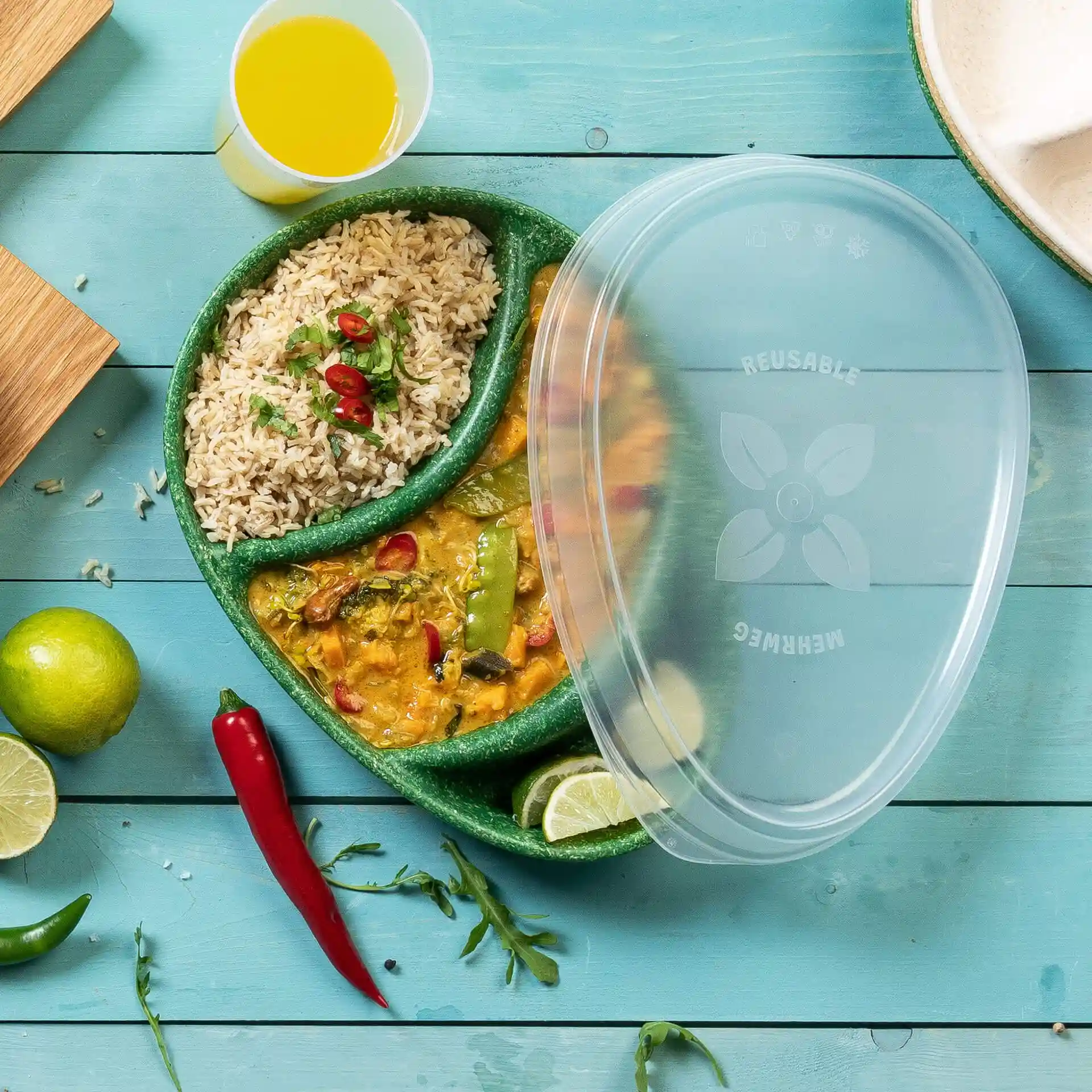 Transparent reusable lid over a divided plate containing rice and curry, with lime and chili peppers on a blue wooden surface.