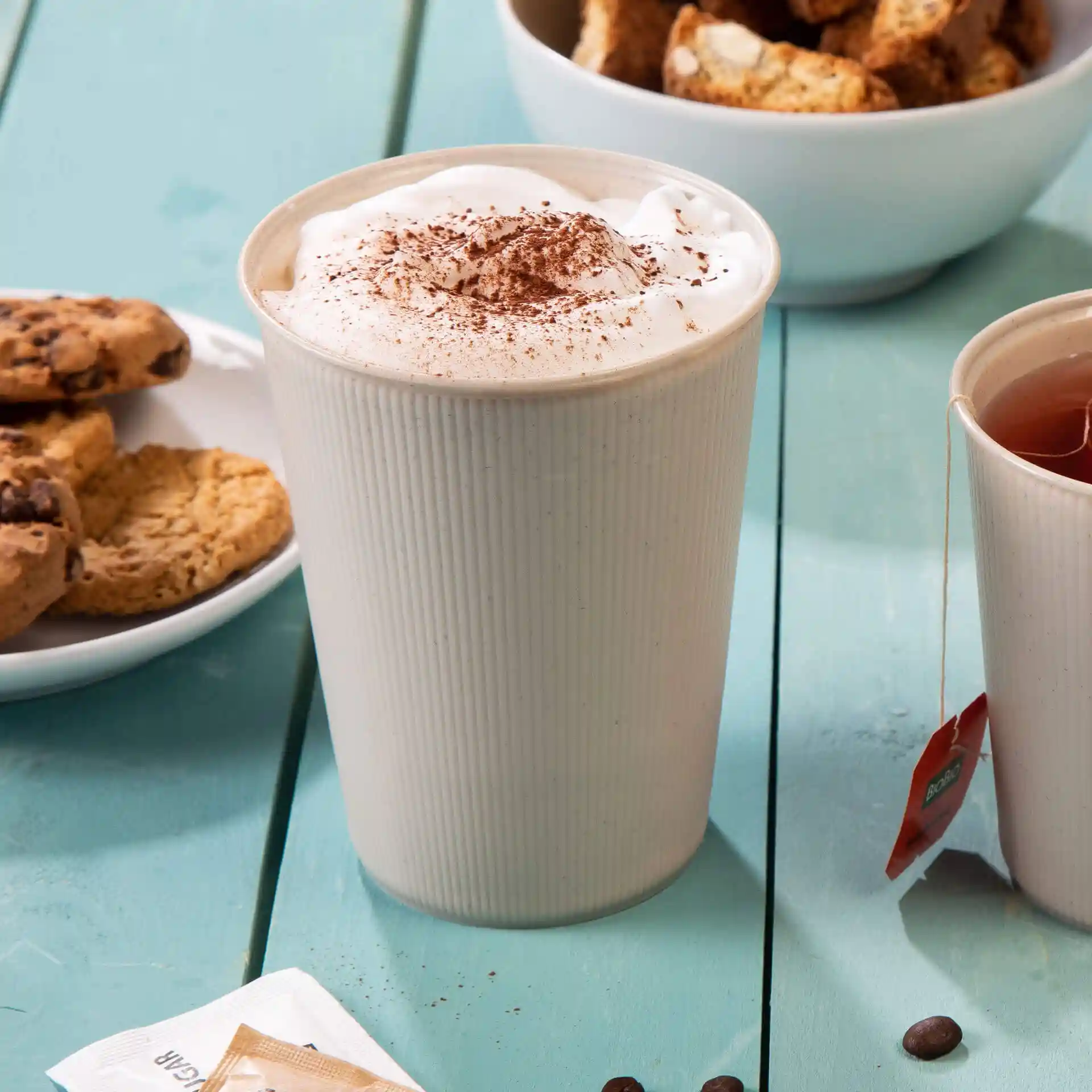 Cream white ribbed reusable cup filled with whipped cream and cocoa, with cookies and tea nearby on a blue table.