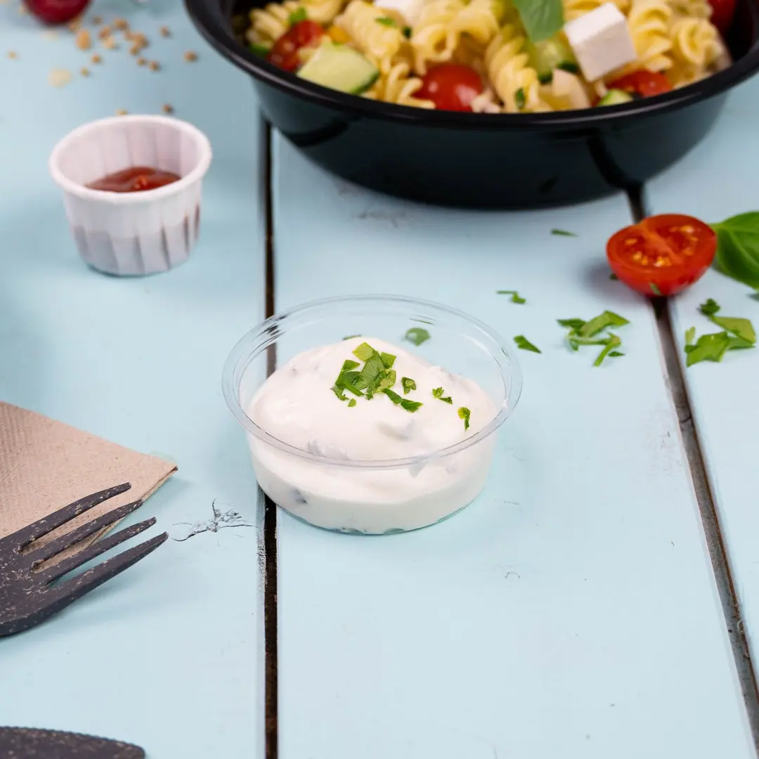 Clear plastic portion cup with creamy sauce topped with chopped herbs, placed on a light blue wooden surface.