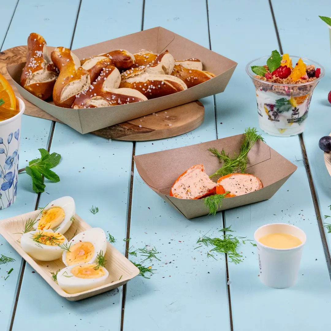 Assorted snacks including pretzels, boiled eggs, yogurt parfait, and a small cup of sauce on a blue wooden surface.