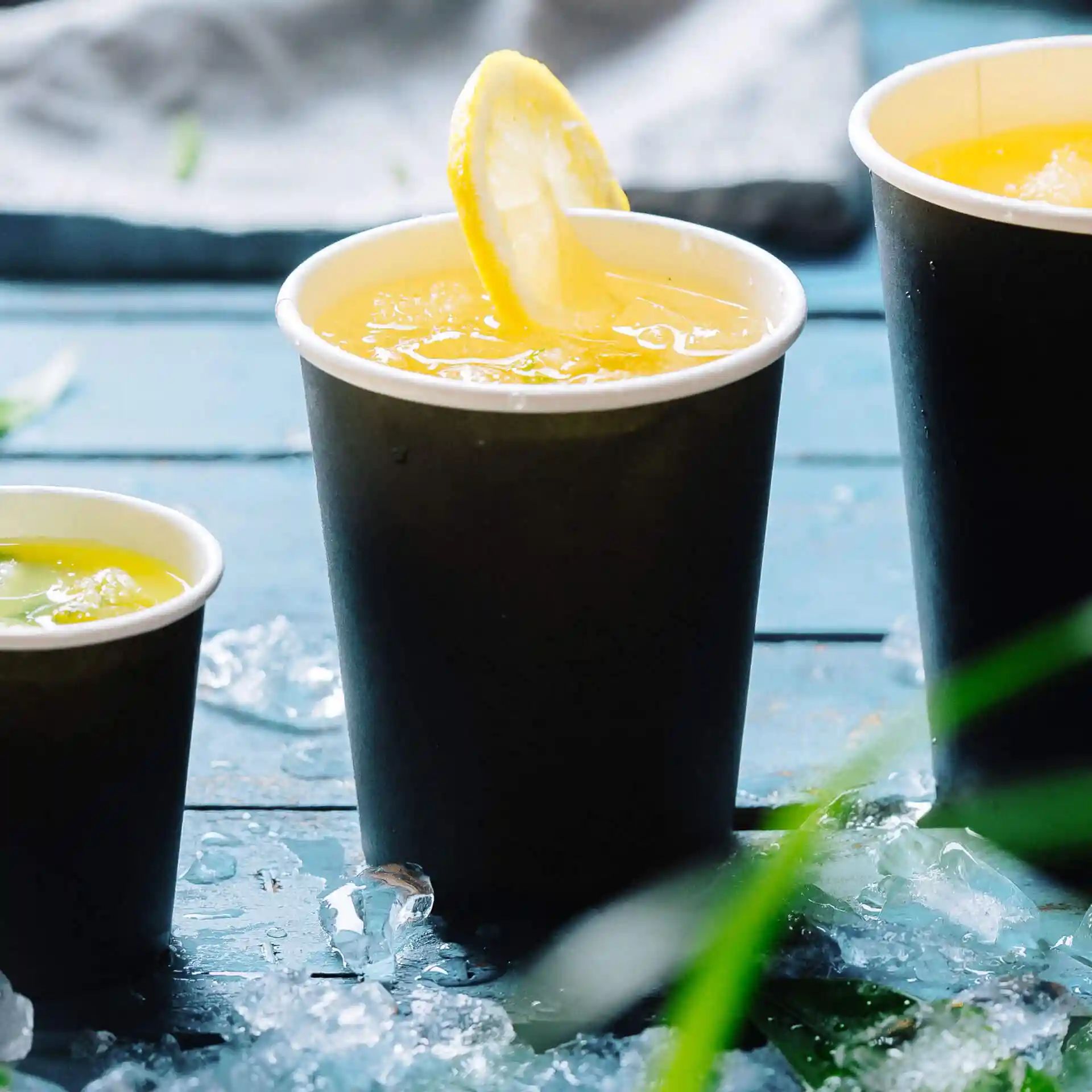 Three black paper cups filled with drinks, one topped with a lemon slice, placed on a blue wooden surface with ice.