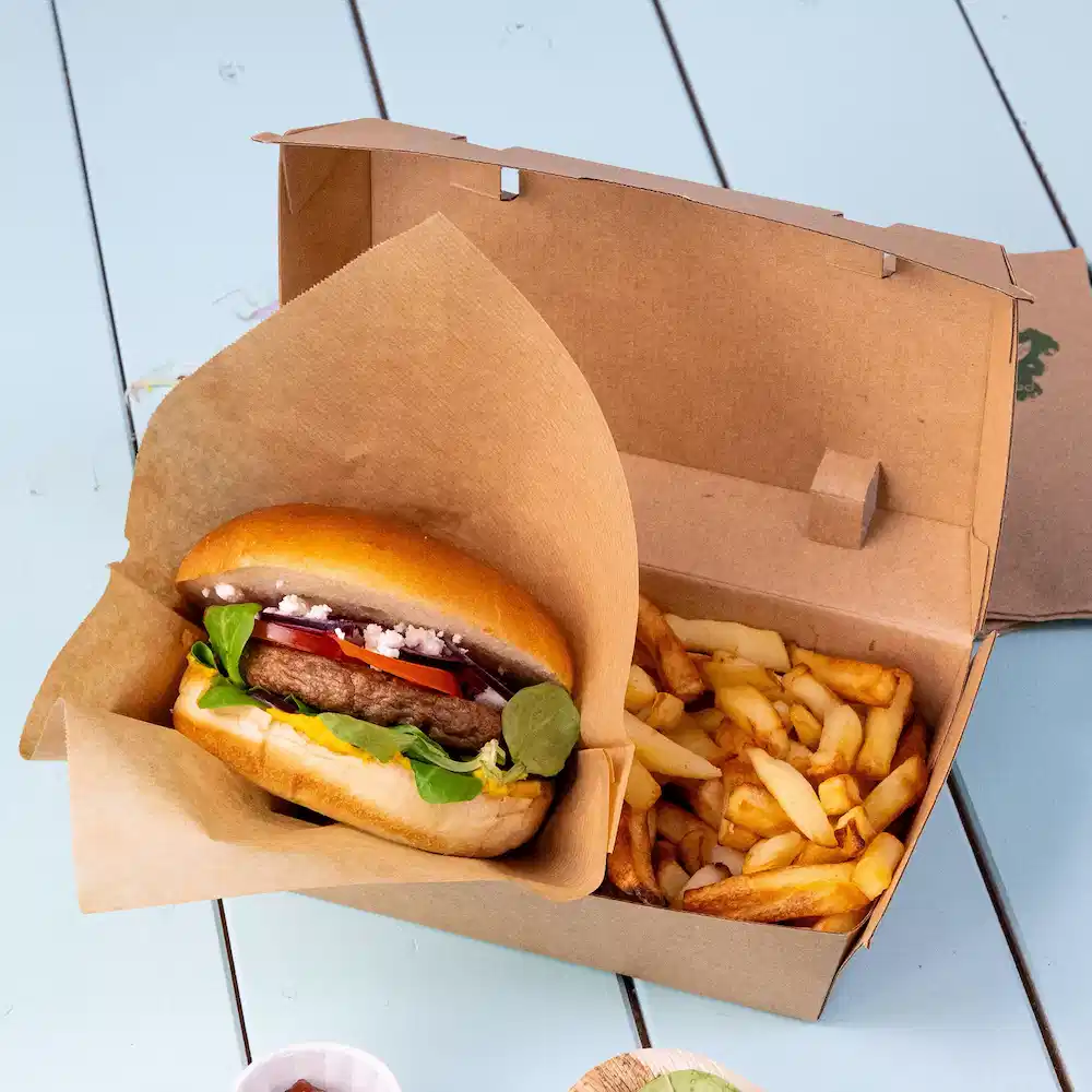 Burger with toppings placed in a takeout box alongside a serving of French fries.