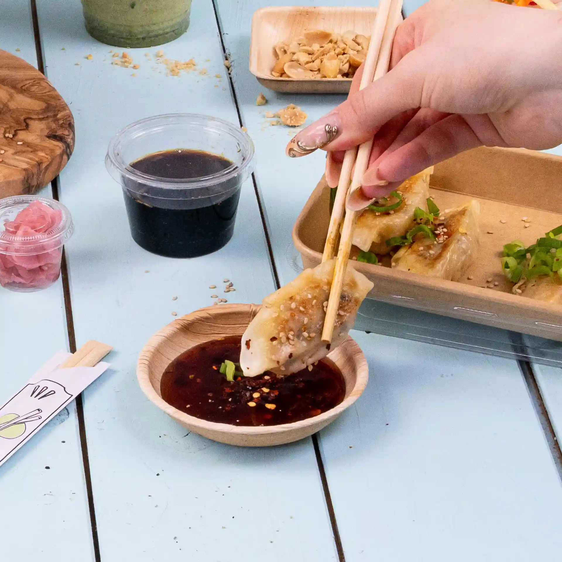 A hand holding chopsticks dips a dumpling into a round palm leaf bowl filled with dark sauce.