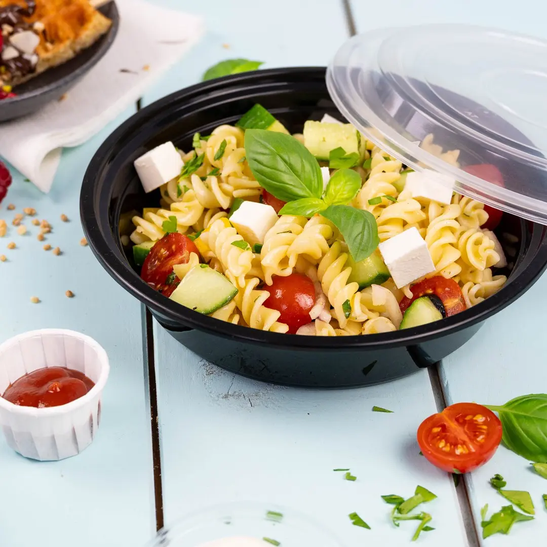 Black bowl with pasta salad containing rotini, cherry tomatoes, cucumber, feta cheese, and basil, with a side of ketchup.