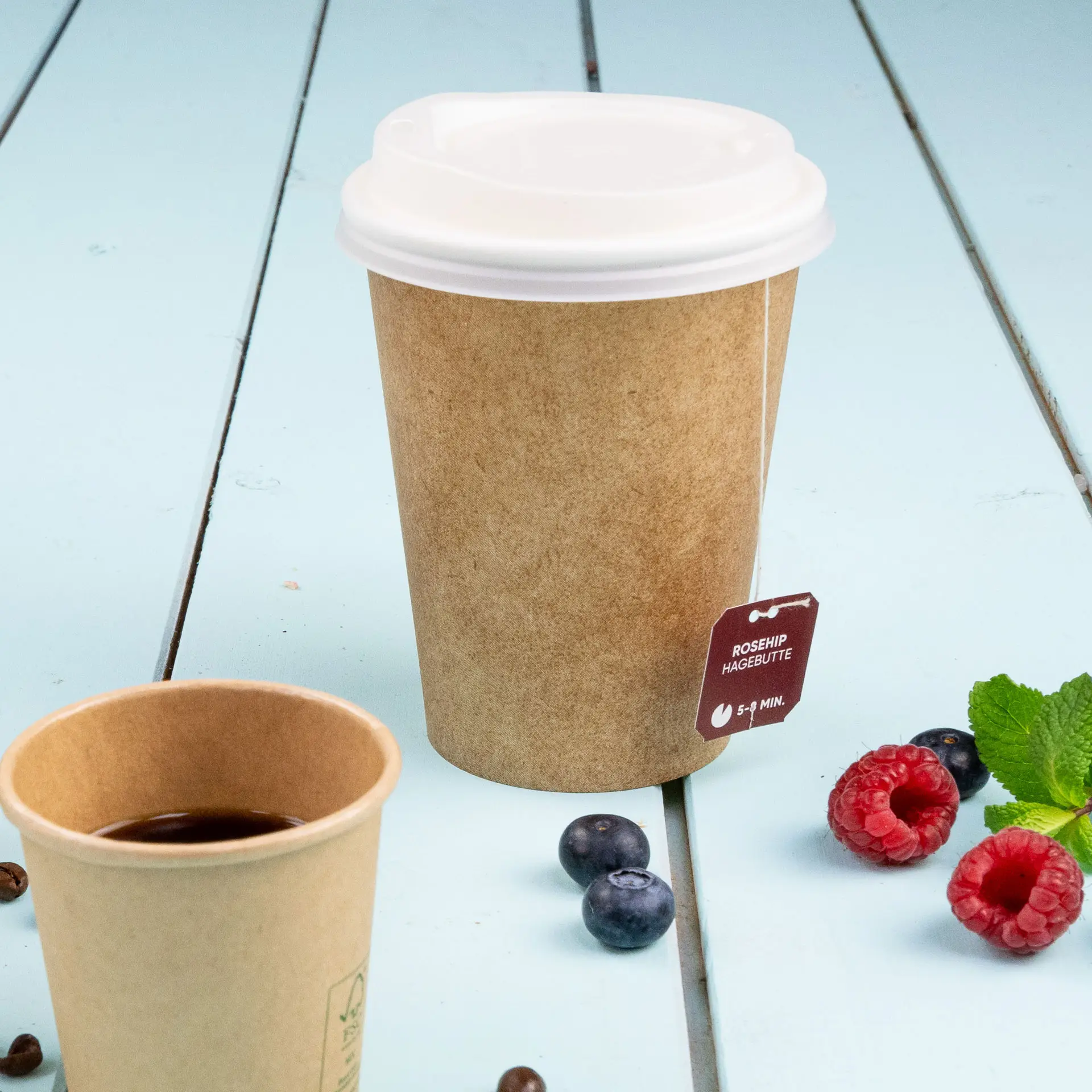 Single-wall brown paper cup with a white lid, featuring a tea bag tag, placed beside a smaller cup and scattered berries.
