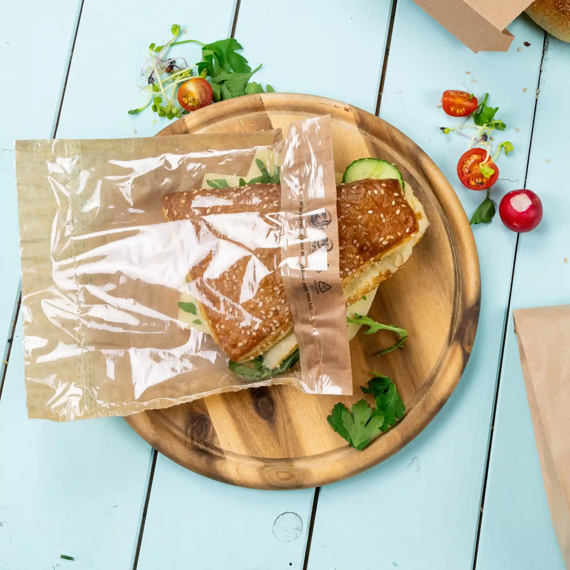 Transparent snack bag with a sesame seed sandwich on a wooden plate, surrounded by fresh herbs and vegetables.