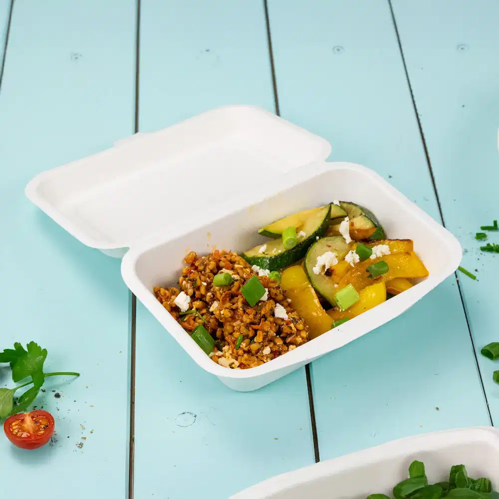 White food container with a mix of cooked grains, zucchini, peppers, and topped with green onions and crumbled cheese.
