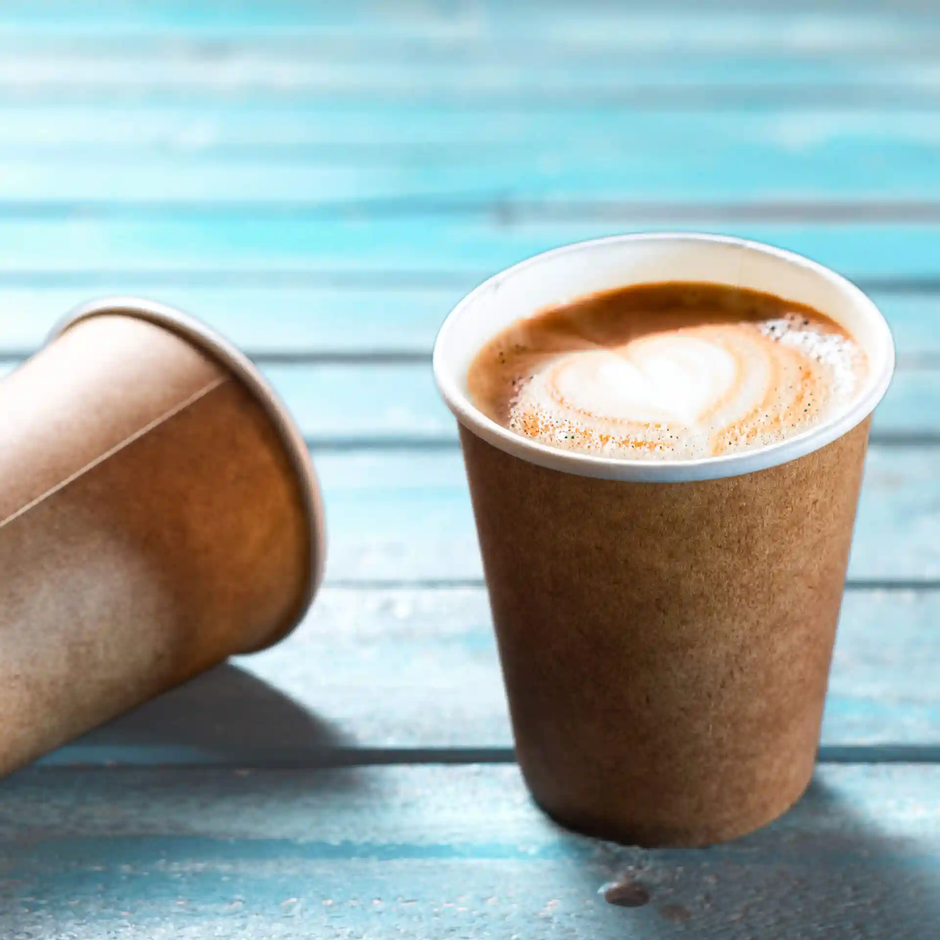 Brown paper cup with a white inner lining, filled with coffee and a heart-shaped latte art, on a blue wooden surface.