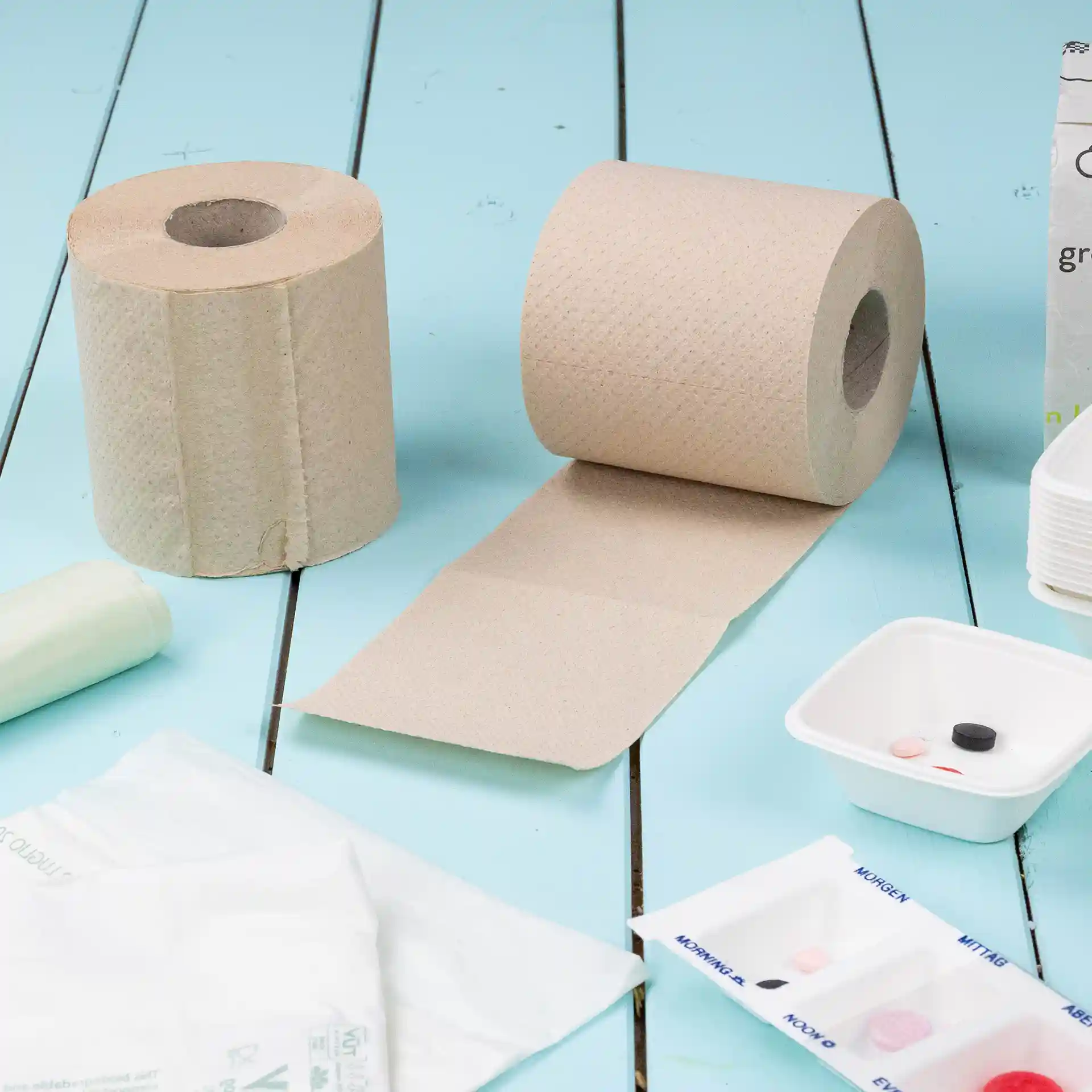 Two rolls of brown toilet paper made from recycled paper, 2-ply, with one roll partially unrolled on a blue surface.