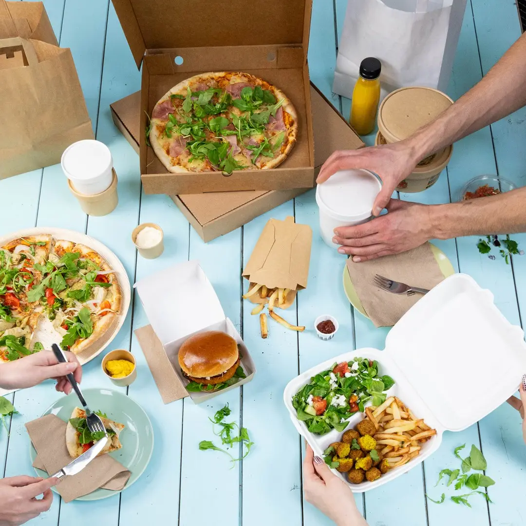 Table set with various takeaway food items including pizzas, a burger, fries, salad, and drinks in containers.